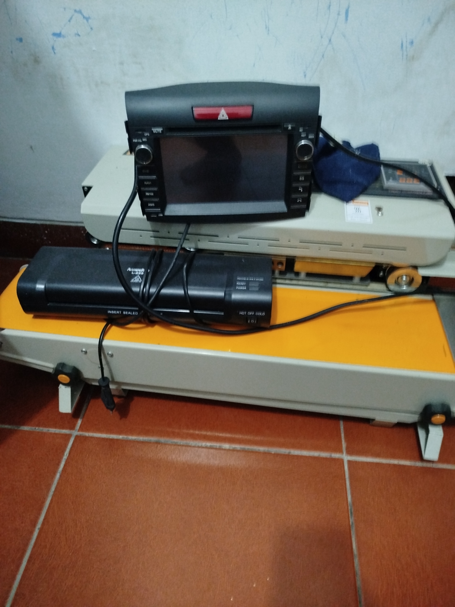 The image shows a car stereo and a laminating machine placed on a table in a room with tiled flooring and a textured wall. The car stereo, a black unit with a screen and various buttons, is positioned on top of the laminating machine. The laminating machine is also black with an orange platform and some visible controls and wires. A dark blue cloth is draped over the side of the car stereo. The room appears to be an indoor space, possibly a workshop or storage area, with the floor tiled in a reddish-brown color and the wall painted a light color with some blue marks. The overall scene is static, with no visible people or ongoing actions.