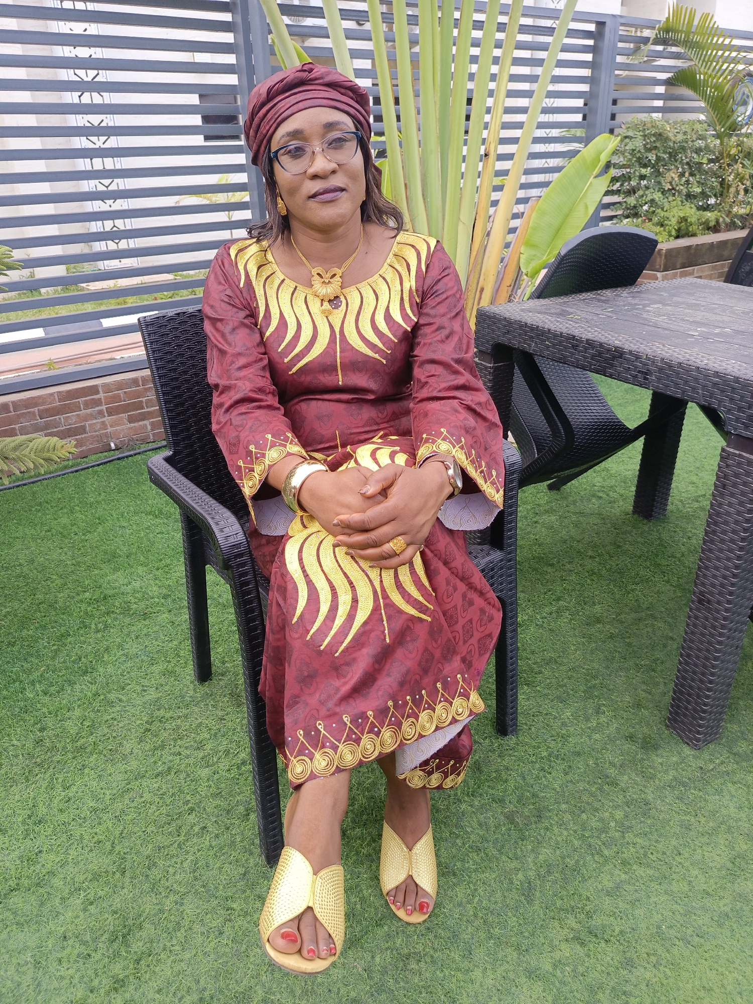 A woman of possibly middle age is seated outdoors, looking directly at the camera with a calm and composed expression. She is the central subject of the image, appearing to pose for a portrait in an outdoor patio or garden setting in Akwanga, Nigeria.
The woman is dressed in a vibrant maroon or deep red traditional garment, likely a dress or tunic, richly decorated with intricate gold embroidery. The gold patterns are prominently featured around the neckline, along the sleeves, and near the hem of the dress. She wears a matching maroon headwrap, carefully styled. Her accessories include dark-framed eyeglasses, a large gold necklace with an ornate pendant, gold earrings, gold bangles on her right wrist, a gold watch on her left, and several gold rings on her fingers. Her feet are clad in stylish gold open-toed sandals, and her toenails are painted red. Her hands are clasped neatly in her lap.
She is seated on a black chair made of woven plastic material. To her right, a matching black woven plastic table and another chair are visible, suggesting an outdoor dining or lounge area. The ground is covered with bright green artificial turf, giving the space a manicured appearance. Behind her and to her right, large, tall green plants, possibly a species of palm or traveler's palm, add to the natural ambiance. Further back, a modern grey fence with horizontal slats and some white decorative patterns can be seen. The overall lighting suggests it is daytime. There is no visible text in the image.