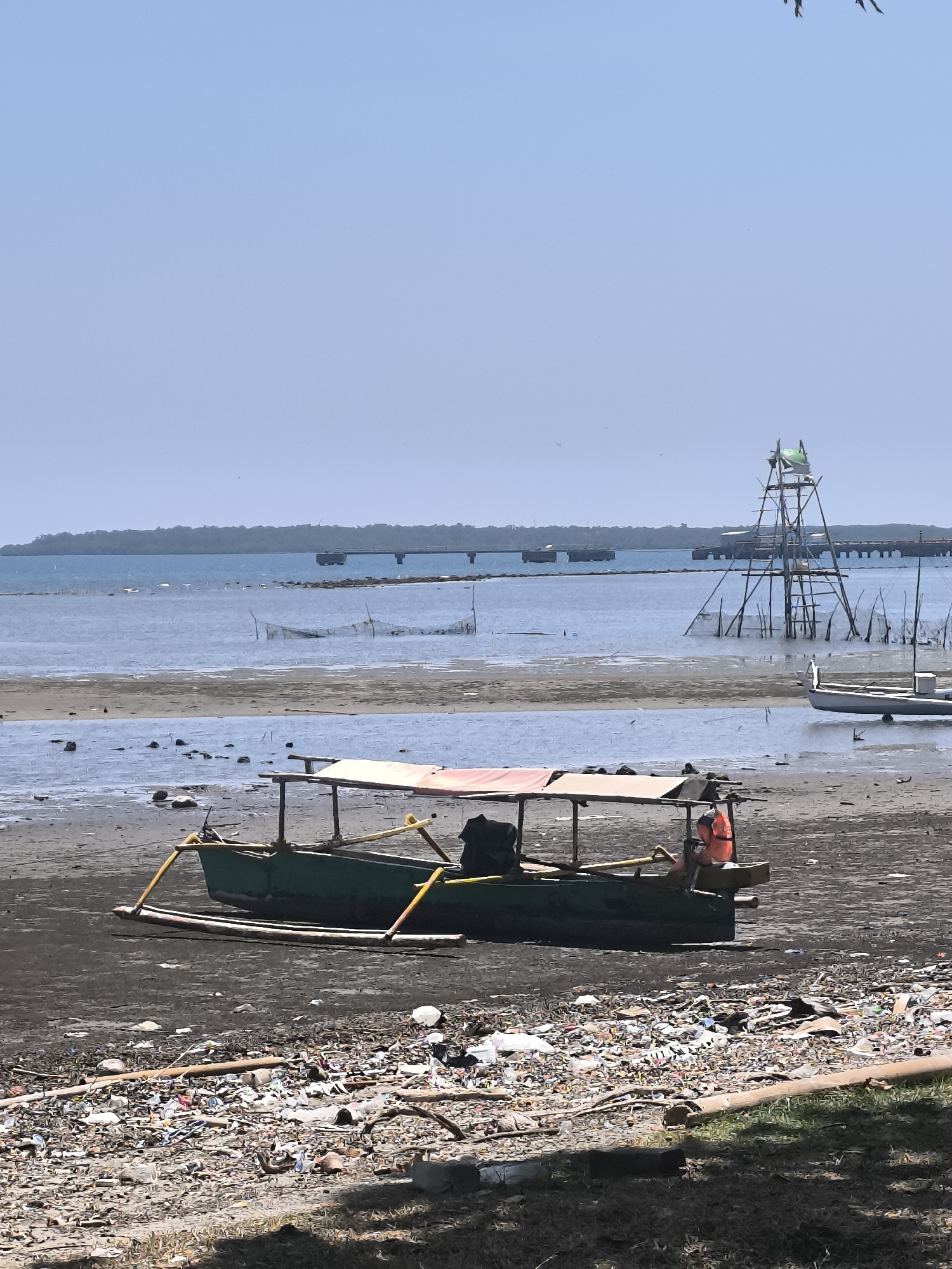 This image depicts a shoreline in Barru, Indonesia, characterized by a significant amount of litter strewn across the dark sand. In the foreground, a traditional green fishing boat with a canopy is partially visible, resting on the shore. An orange life vest is placed on the back of the boat. The water's edge is close, with some debris floating near the boat. In the middle ground, the shallow water reveals the muddy bottom and scattered rocks. Further out, the sea extends towards a distant, hazy coastline with a long pier or bridge structure and a wooden observation tower with a green flag. The sky is clear and light blue. The overall scene suggests a polluted coastal environment.
