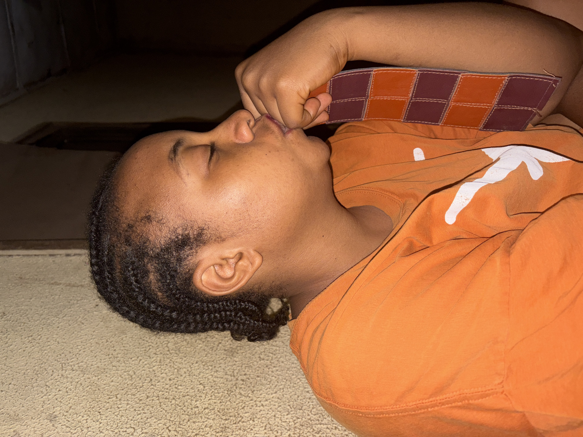 A young person with dark skin and braided hair is captured lying on their side on a light-colored, textured floor in a dimly lit indoor setting in Jos, Nigeria. The individual is seen in profile, with their eyes closed, suggesting they are resting or sleeping. They are wearing a bright orange t-shirt featuring a prominent white graphic that resembles the "Hook 'em Horns" symbol, commonly associated with the University of Texas. One hand is raised and positioned near their mouth, holding a small, rectangular object with a checkered pattern of alternating orange and brown squares, possibly as a comfort item. The background is very dark, indicating low light conditions, likely at night. The person's relaxed posture and closed eyes convey a sense of peaceful slumber or quiet repose.