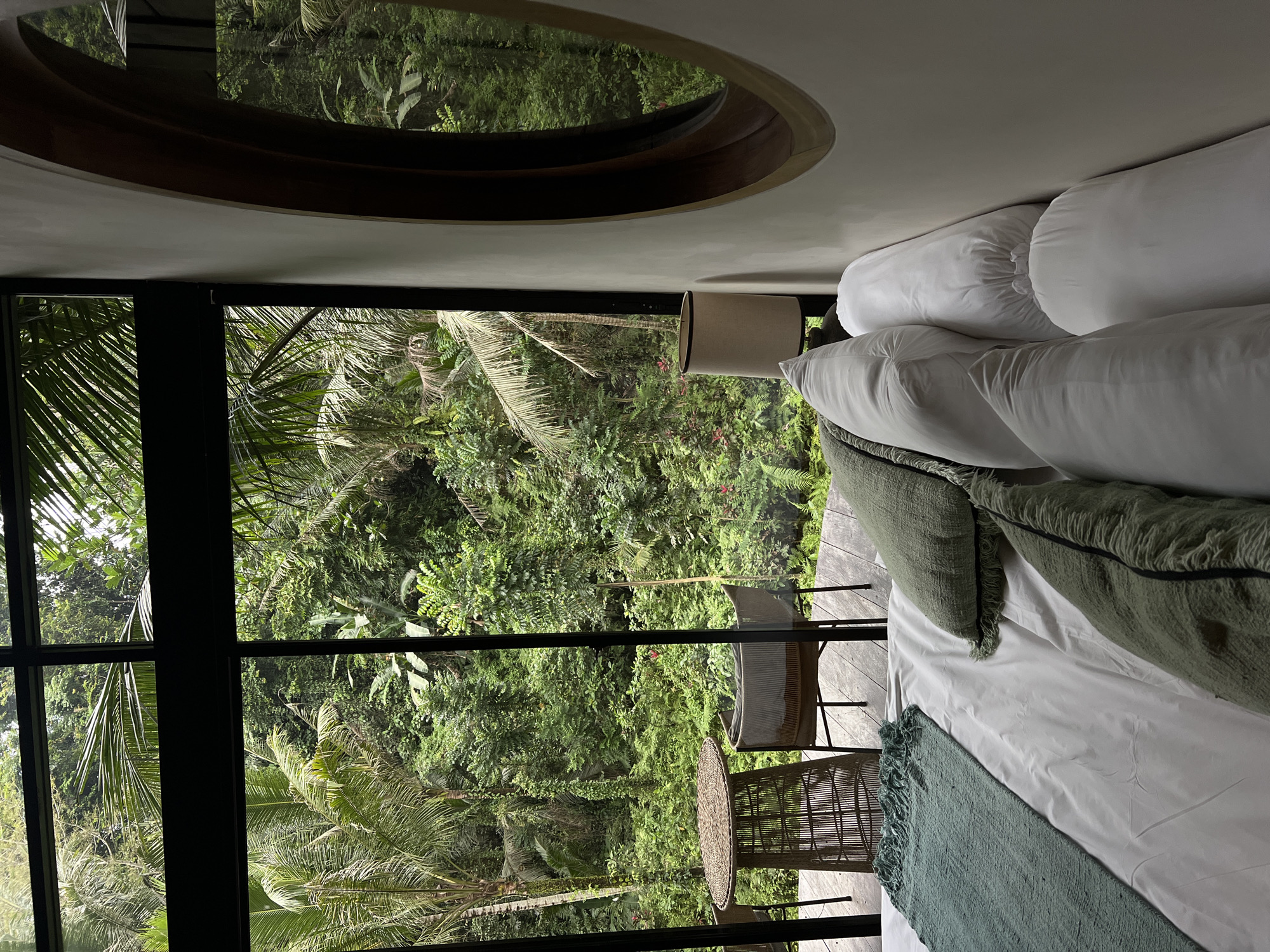 This media file shows a bedroom with a large window overlooking a lush, green jungle landscape. The room is furnished with a bed with white sheets and several pillows, two of which are green and have a fringed texture. A teal-colored throw blanket is draped across the foot of the bed. Next to the bed is a bedside table with a lamp on it. Outside the window, there is a small outdoor seating area with a round, woven table and two chairs. Beyond the seating area, the dense jungle foliage is visible, with palm trees and other tropical plants. The scene is brightly lit, suggesting it is daytime. The overall impression is one of tranquility and immersion in nature. The location is the Ubud District in Indonesia, known for its rich cultural heritage and tropical rainforests. There is no visible text in the image.