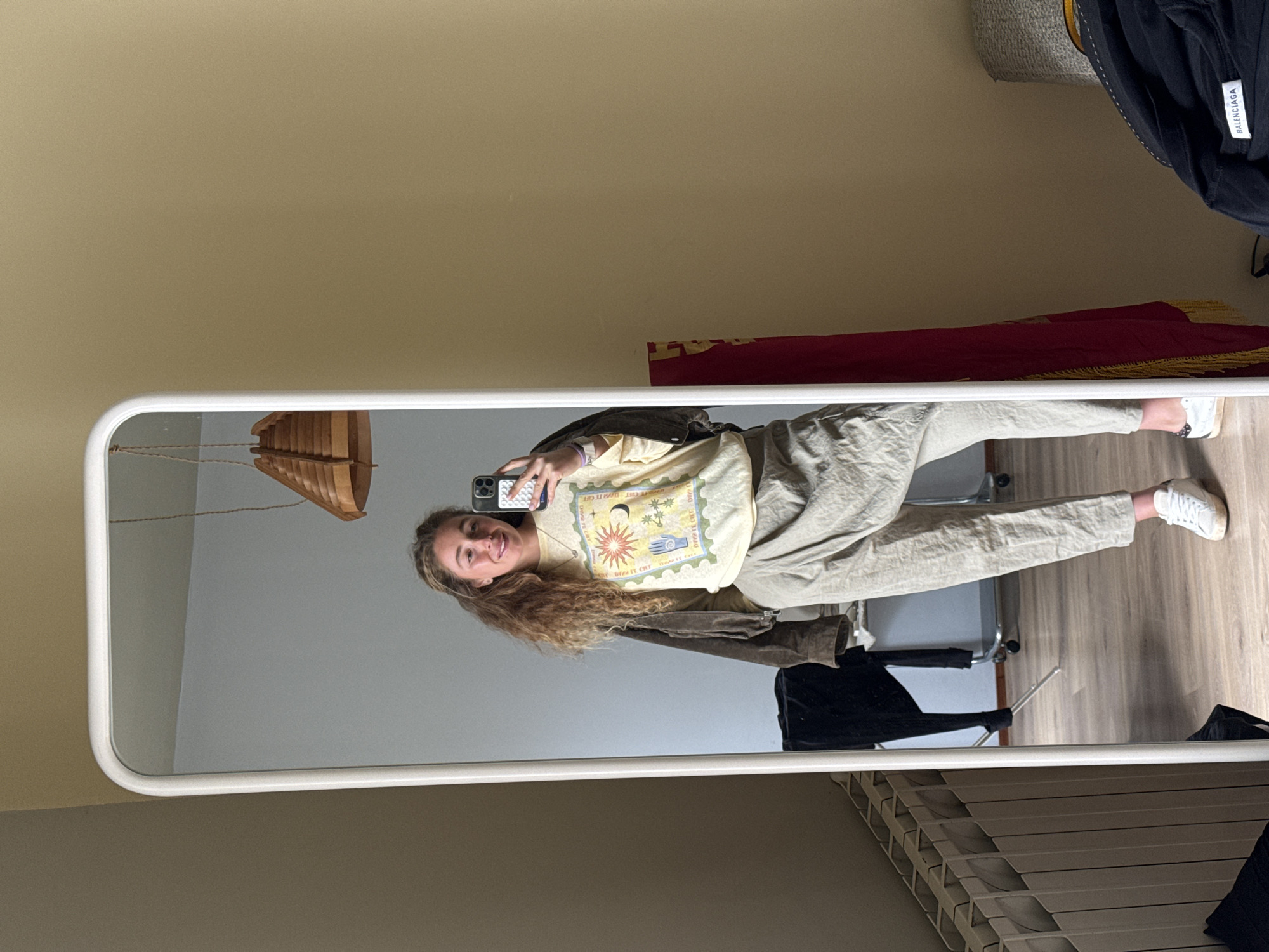 A young woman stands in front of a tall, rectangular mirror with a white frame, taking a selfie. She is wearing a light yellow t-shirt with a graphic design, light beige pants, a brown jacket, and white sneakers. She is holding a smartphone with a black and white case. The mirror reflects the interior of a room with light beige walls. A wooden pendant light hangs from the ceiling. Behind the woman in the mirror are a silver chair, a black garment on a hanger, and a white slatted closet door. On a shelf above the mirror's reflection is a red and gold embroidered fabric, as well as a black jacket. The room has a wooden floor. There is no evidence to suggest the location is Madrid, Spain.