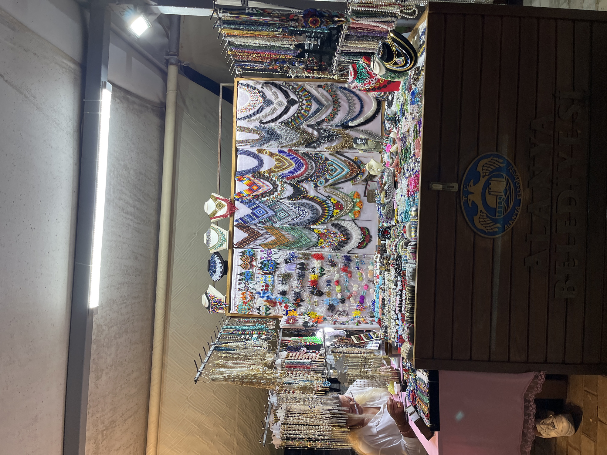 This media file captures an indoor market stall, likely within a bazaar or covered market, showcasing a vibrant array of jewelry and accessories. The scene is illuminated by artificial overhead lighting, including fluorescent strips and a spotlight, suggesting an evening setting or an interior space without natural light.
The primary subjects are the multitude of colorful items for sale. Numerous necklaces, many appearing to be intricately beaded or embroidered with geometric and abstract patterns in a wide spectrum of colors, are prominently displayed on various stands and flat surfaces. Among them are beaded chokers, longer chain necklaces, and those adorned with shells or pearls. Alongside these, various bracelets, rings, and hair accessories such as clips and hair ties are neatly arranged on racks and display boards, contributing to the rich visual tapestry of the stall.
The stall itself features a dark brown, vertically paneled wooden counter or display cabinet. Affixed to this wooden structure is a circular blue and gold emblem. This emblem clearly displays the text "T.C. ALANYA BELEDİYESİ," which translates to "Republic of Turkey Alanya Municipality." The same text, "ALANYA BELEDİYESİ," is also carved or painted vertically onto the wooden stall, serving as a strong location cue.
At least one person is visible behind the counter, partially obscured, with light-colored hair and wearing a white shirt, possibly a vendor or a shopper examining items. Another person, wearing a face mask and dark clothing, is partially seen in the lower right, also interacting with the merchandise. The activity taking place is typical of a market, involving the display, browsing, and potential sale of goods. The walls of the market area are light-colored, possibly concrete or painted, showing some textured wear.