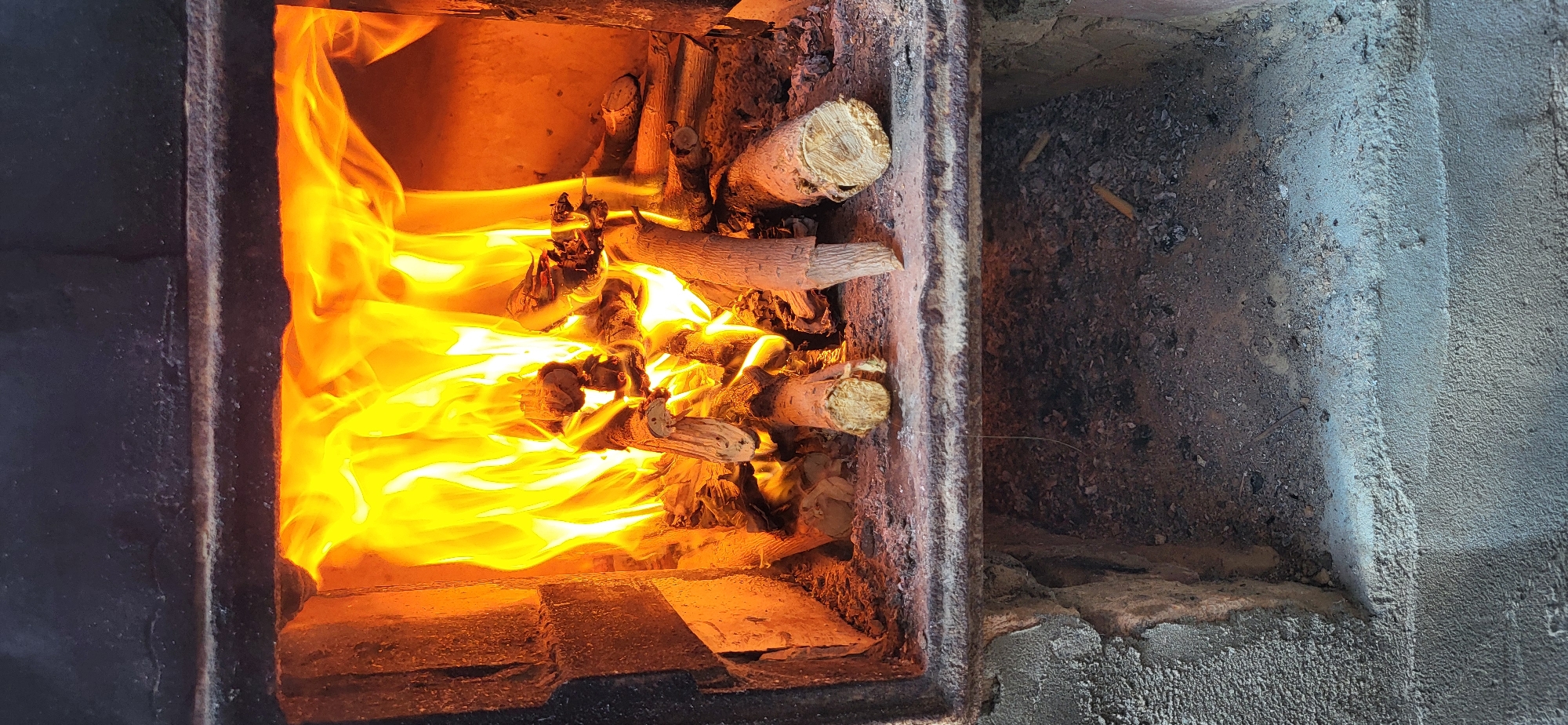 An interior view of a lit fireplace reveals a roaring fire with vibrant orange and yellow flames consuming several pieces of split firewood. The logs, some with visible bark and cut ends, are arranged on a grate. The fireplace is constructed of brick and metal, with a dark metal frame surrounding the opening. Below the grate, a lower compartment of the fireplace is visible, filled with ash and debris. The scene appears to be a domestic setting within the city of Taldykorgan, Kazakhstan, possibly a residential home or a communal area with a fireplace. There are no people or animals present in the image, and the focus is solely on the burning fire and the fireplace structure.
