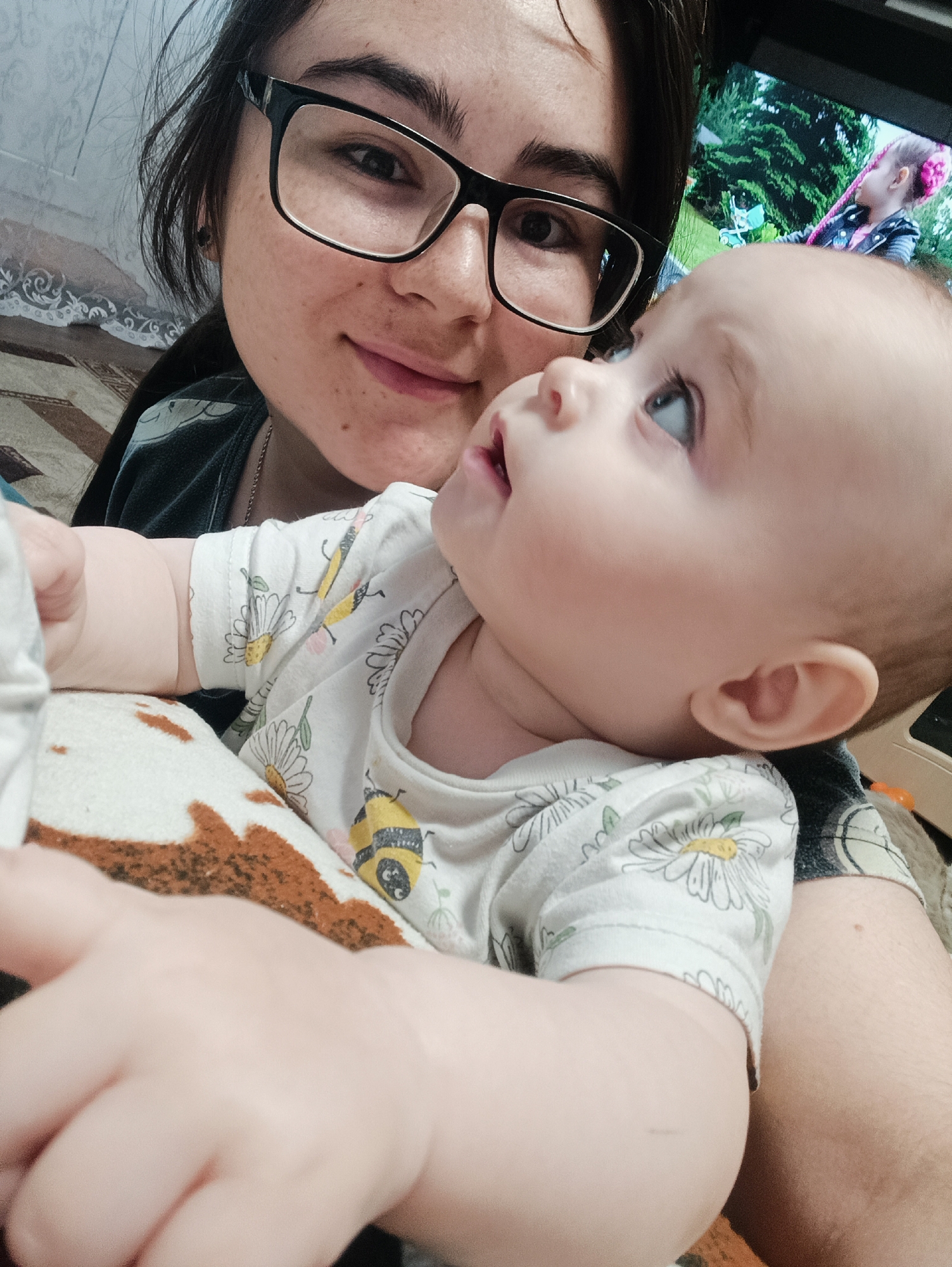 The image presents a close-up, intimate portrait of a young woman and a baby indoors. The woman, likely the mother, is positioned in the foreground on the left, looking down and to the right with a gentle smile. She has dark hair, wears black-framed glasses, and a dark-colored top, with a thin necklace chain visible.
The baby, an infant, is held close to the woman, situated in front of her. The baby looks upwards and to the right with a curious expression, its mouth slightly open. The baby wears a white short-sleeved onesie decorated with yellow bumblebees and white daisies. Its plump left arm and hand are prominent in the lower foreground.
The background reveals elements of a home environment. To the far left, a patterned wallpaper or fabric is partially visible. On the right, a television screen displays a colorful scene from what appears to be a children's program, showing a character with pink hair in an outdoor, green setting. A brown and white patterned blanket or soft furnishing is visible beneath the baby.
The activity captured is an affectionate interaction between the woman and the baby, possibly a selfie or a close-up photo taken by the woman. The overall mood is tender and warm. The lighting suggests an indoor setting, and there is no discernible text visible in the image.
