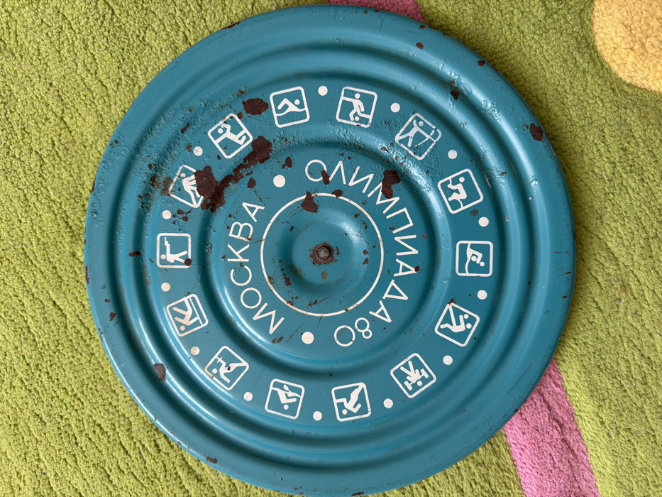 The image displays a circular, teal-painted metal disc resting on a textured rug. The disc is significantly worn, exhibiting numerous scratches, paint chips, and irregular patches of dark brown rust, particularly concentrated on the right side and near its center. Concentric ridges are visible across the disc's surface. At the center, a dark metal rivet or screw is present. White Cyrillic text is arranged in a circular pattern around the center: "МОСКВА" (Moscow) at the top and "ОЛИМПИАДА 80" (Olympiad 80) at the bottom, partially mirrored. Surrounding this text is an outer ring of white pictograms, each depicting an Olympic sport within a square, separated by small white dots. Examples of visible pictograms include shooting, running, weightlifting, swimming, and others. The background is a lime green carpet or rug with a visible pink stripe running diagonally in the bottom left portion and a circular yellow patch in the bottom right. No individuals or active interactions are present. The specific location within Chaikovsky, Russia, cannot be determined from the visual information provided.