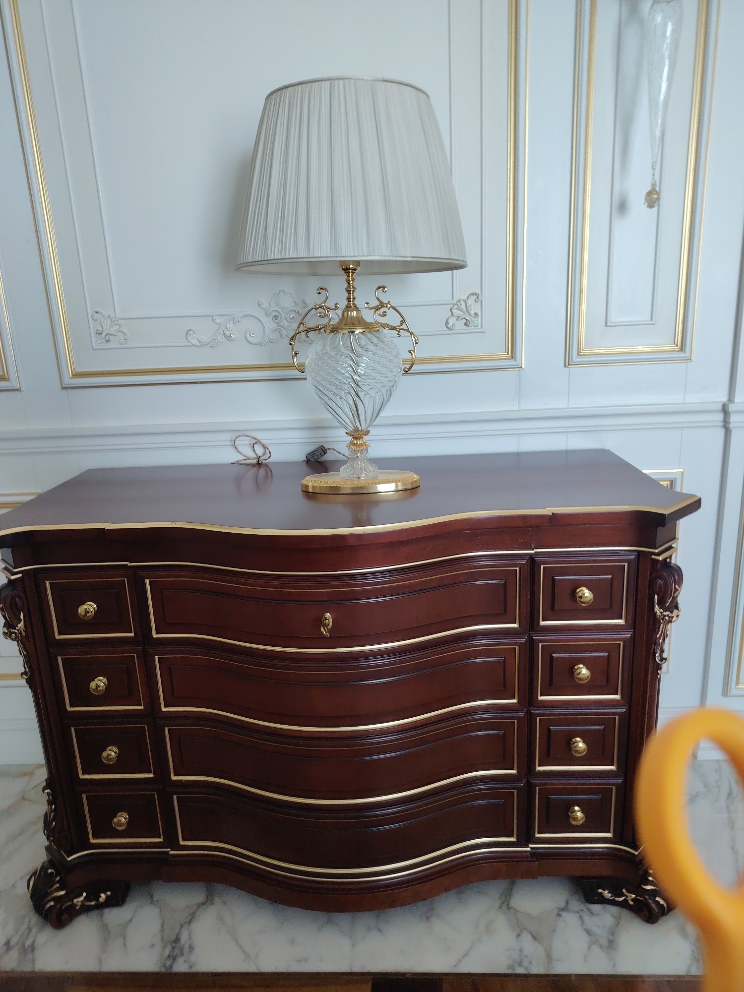 The image shows a close-up view of a wooden dresser with gold trim, a table lamp with a pleated shade and a glass base, and decorative molding on a white wall in the background. The dresser is dark wood with multiple drawers, each featuring gold hardware and trim. The table lamp is placed on top of the dresser, and its base is ornate glass with gold accents. The wall behind the dresser is decorated with white paneling and gold trim, creating a formal and luxurious aesthetic. The floor appears to be marble. There is no indication of the location being in Konigil, Uzbekistan, as the image lacks any discernible Uzbek architectural or cultural features.