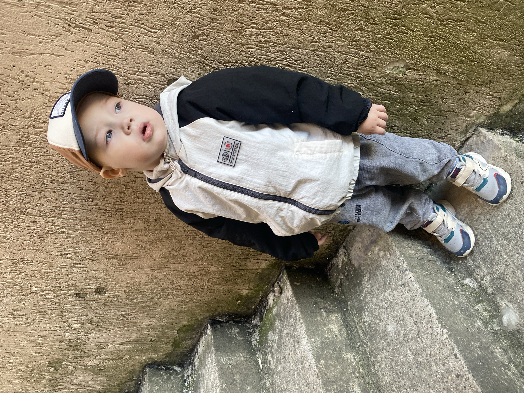 A young child is depicted outdoors, standing on concrete steps with a textured concrete wall on their left. The child, a toddler with fair skin and light blue eyes, looks upwards with a slightly open mouth, conveying curiosity or wonder. They are dressed in a multi-toned jacket, featuring a black upper section and off-white/light beige lower half with a black zipper down the front. Grey sweatpants are worn on their legs, and multicolored sneakers (grey, white, blue, green, and red accents) are on their feet. A baseball cap, primarily beige and dark blue with a brown brim, sits on their head, featuring a small, mostly illegible patch on the front, possibly reading "SHARK".
On the off-white section of the jacket, a rectangular patch displays text including "OUT OF FRAME", "GO IN", "GO OUTSIDE", alongside a small graphic of a globe. Text is also visible on the left leg of the sweatpants, appearing to read "TRAILDEK RIVKLE".
The surrounding environment is rugged; the concrete wall and steps show signs of age, with patches of dark moss or algae on their surfaces, suggesting a damp or shaded outdoor area. The lighting is bright and even, indicating daytime, possibly under an overcast sky or in a shadowed location. The image captures a candid moment of the child observing something above them.