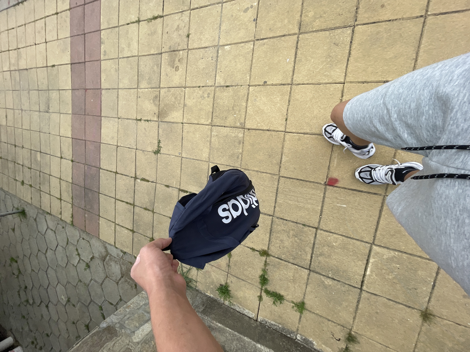 The image, taken from a high-angle perspective, captures an outdoor scene in Zlatograd, Bulgaria. The primary subject is a person, visible from their legs, feet, and an outstretched arm and hand. The person is wearing light grey sweat shorts or track pants and a pair of black and white athletic shoes with white laces. One of the person's hands is holding a navy blue or dark blue sling bag or fanny pack, which features the word "adidas" printed upside down in white lettering.
The setting appears to be a paved area, likely a sidewalk or pathway. The ground is covered with square, light beige/yellowish paving stones, with a central strip of reddish-brown rectangular tiles running through them. Small patches of weeds and grass are growing in the cracks between the tiles. To the lower left, there is a drop-off revealing a concrete retaining wall constructed from hexagonal blocks, indicating the presence of a slope or a channel below the paved surface. A metal pipe is also visible extending from the ground near the top of this wall. The lighting suggests it is daytime, possibly overcast, as there are no harsh shadows. No specific activity beyond standing and holding the bag is discernible.