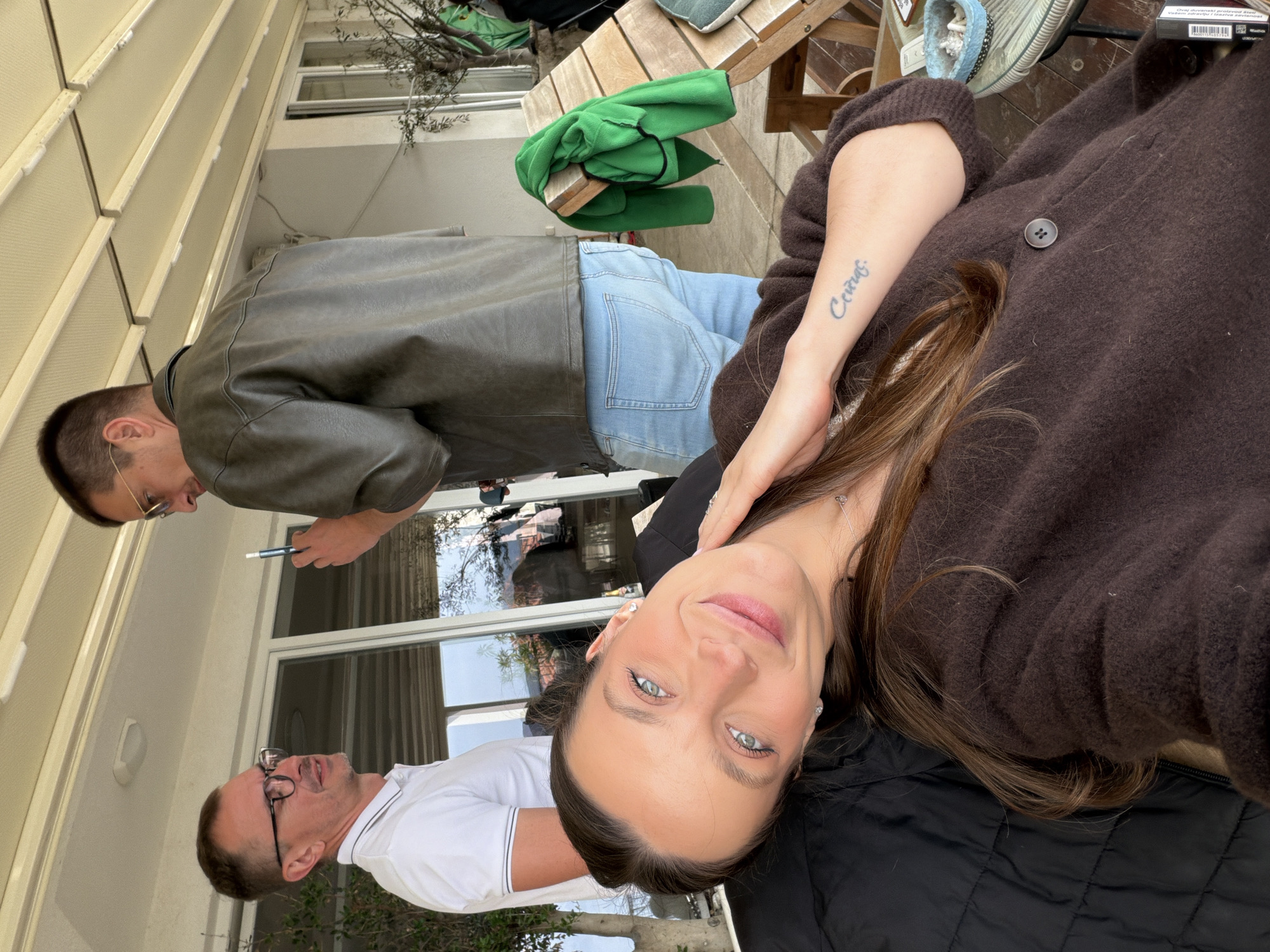 The image depicts a woman in the foreground, wearing a brown cardigan, with a tattoo visible on her arm. Behind her are two men; one in a white collared shirt and glasses, and the other in a leather jacket. They appear to be in an outdoor setting, possibly a patio or backyard, with a building visible in the background and foliage indicating a garden or natural environment. The image lacks sufficient detail to conclusively determine if the location is in the city of Trebesin, Montenegro, or to infer any activities or interactions beyond their mere presence in the frame.