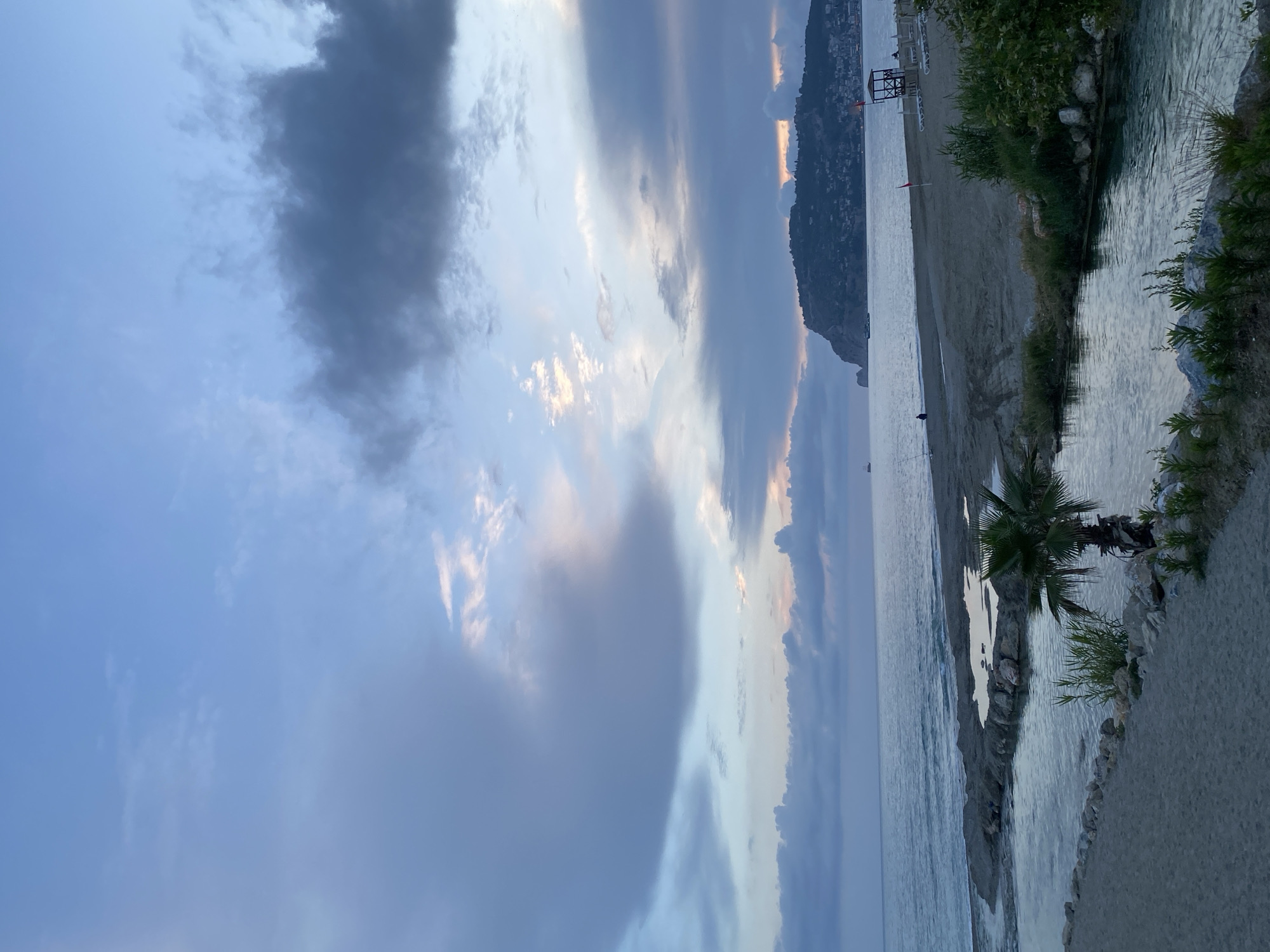 The image captures a tranquil beach scene in Alanya, Turkey. The sky is dominated by dramatic clouds with varying shades of gray and hints of sunlight peeking through. Below, the calm sea stretches towards the horizon, reflecting the sky's colors. A long, sandy beach curves along the coast, ending at a promontory topped with buildings.
On the right, a small river or stream flows into the sea. Vegetation lines the banks of the water, including palm trees and other green plants. A lifeguard tower is visible in the distance along the beach. The scene is quiet, with no distinct human activity immediately apparent, though there might be some people in the distance on the beach. The photograph is taken from a slightly elevated perspective.