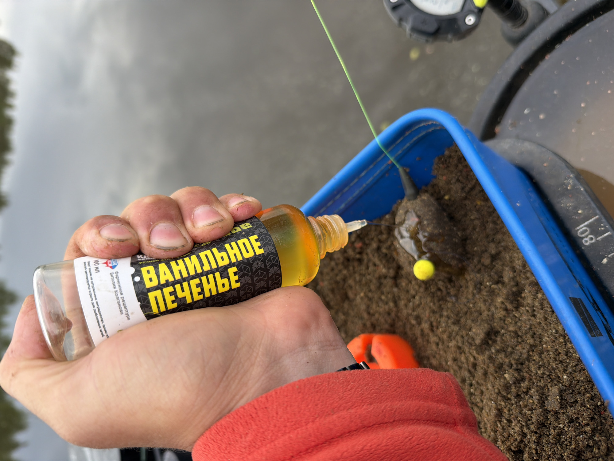 A person is adding "VANILLA COOKIES" liquid attractant to a blue container of bait, likely for fishing. The container appears to be sitting on a fishing seat. The background shows a body of water, possibly a river or lake. The text on the bottle is in Russian, and identifies the attractant as "VANILLA COOKIES" with the recipe by Vitaly Kolganov.