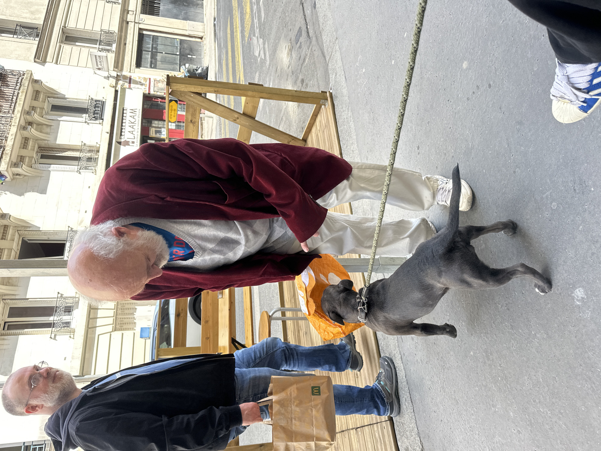 The image depicts a street scene in Paris, France. An elderly man with white hair and a burgundy jacket stands next to a dog on a leash. A younger man in a black jacket and jeans holds a McDonald's bag. The dog is interacting with an orange bag. A restaurant sign is partially visible in the background. A makeshift wooden structure is also present on the sidewalk.