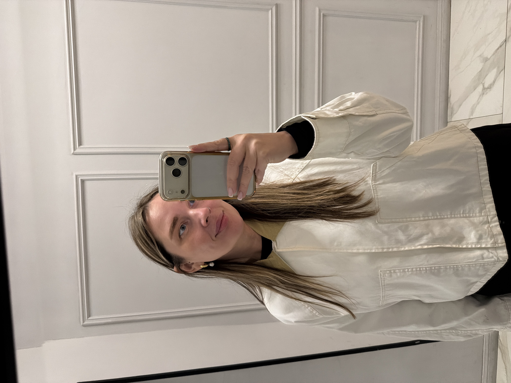 A woman is taking a selfie in front of a mirror. The mirror is in a room with white walls and a marble floor. The woman is wearing a white jacket and black shirt. She has brown hair and blue eyes. She is holding a smartphone in her hand. The location is not specified as Moscow, Russia.