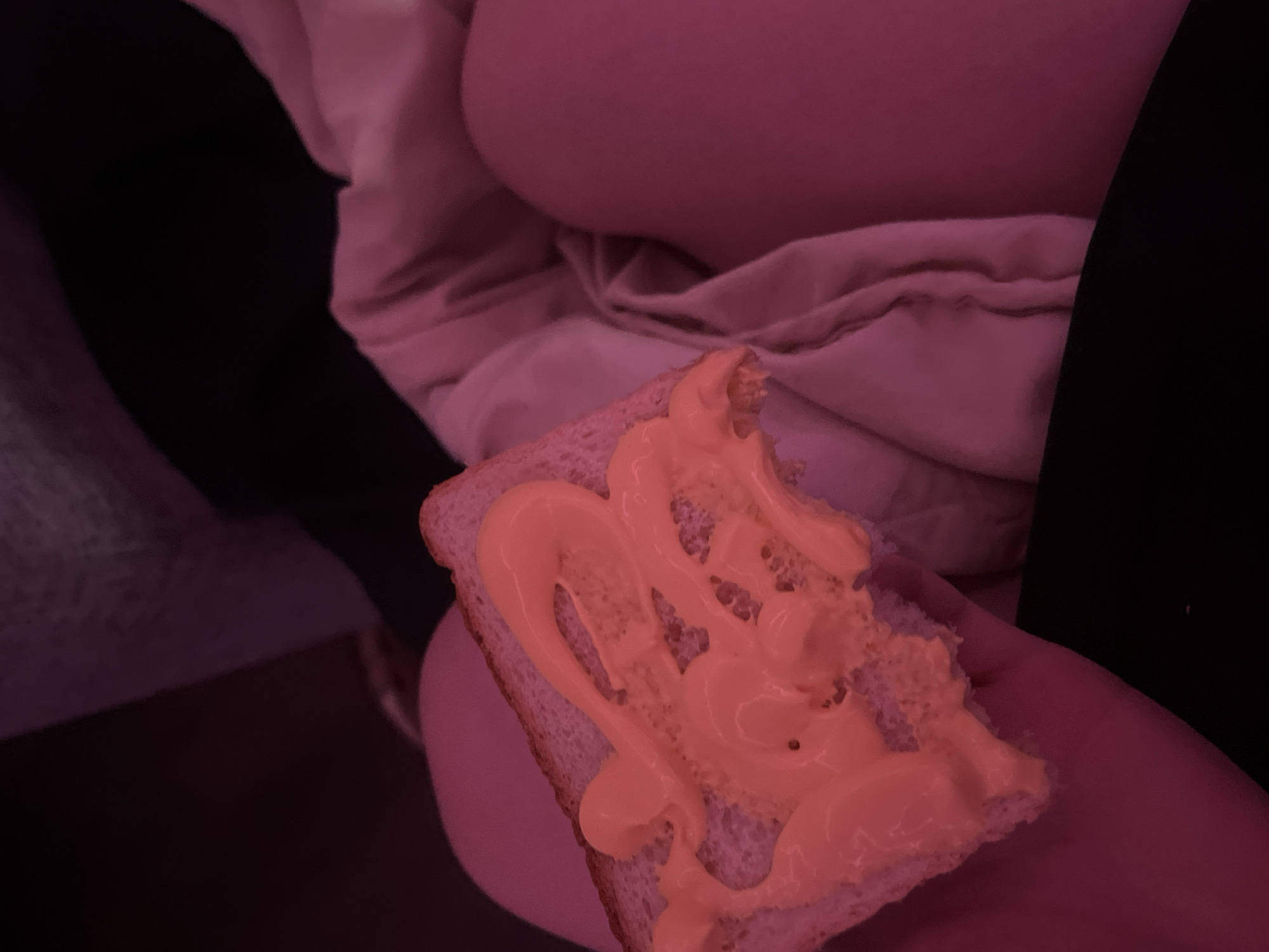 A close-up, low-light indoor photograph captures a hand holding a slice of bread topped with a creamy, orange-yellow spread. The entire scene is bathed in a strong, reddish-pink ambient light, giving all elements a warm, deep hue. The bread appears to be a standard slice of white or light whole wheat bread, with the spread applied in thick, swirling patterns, possibly from a tube or piping bag.
In the blurred background, soft, rumpled fabrics are visible, including a large section of light pinkish-beige material that could be bedding or a blanket, and darker fabric, possibly clothing, on the left. A smooth, rounded form is partially visible behind the bread, which could be part of a person's body or an object covered by fabric. The lighting suggests it is evening or night. No specific location cues, weather details, or visible text are present. The activity depicted is the holding of a prepared snack, likely before consumption.