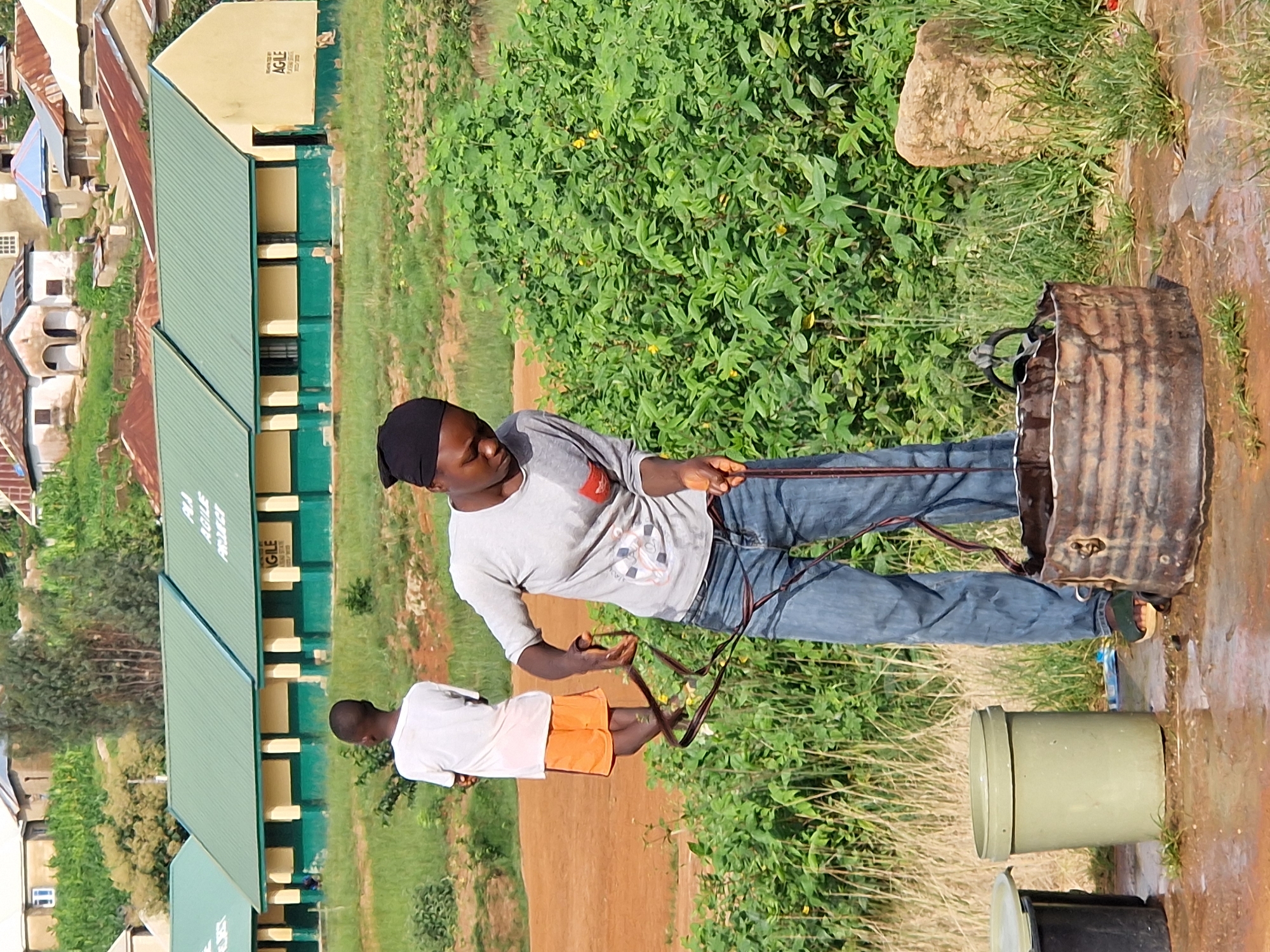This photograph, taken in Kabong, Nigeria, depicts an outdoor scene near a school building with a green roof. In the foreground, a person wearing a black headscarf, a gray t-shirt, and blue jeans is actively using a rope, possibly for drawing water from a metal basin, which appears to be a makeshift well. Next to this basin are two plastic buckets, one light green and the other black, positioned on wet, muddy ground. To the left of these buckets, a young boy in a white t-shirt and orange shorts stands with his back to the camera, looking towards the school. The school building itself has a yellow facade with green trim and signage that reads "AGILE PROJECT." Behind the school, a hilly landscape is visible, dotted with various houses with corrugated iron roofs, suggesting a residential area. The overall environment is rural or semi-rural, with patches of dry grass and cultivated land in the background.