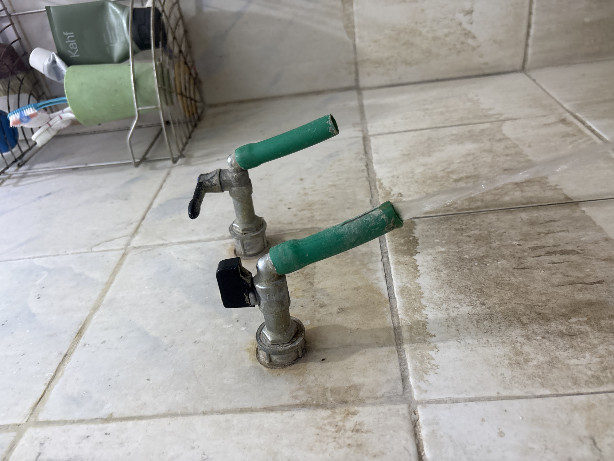 The image shows two water faucets on a tiled wall. Both faucets have green hoses attached and water is running from the right faucet. The tiles on the wall are light-colored and appear dirty with brown stains. In the background, a shelf holds a toothbrush holder with toothbrushes and a tube of Kahf skincare product. The scene appears to be in a bathroom, possibly in Palimanan, Indonesia, given the context.