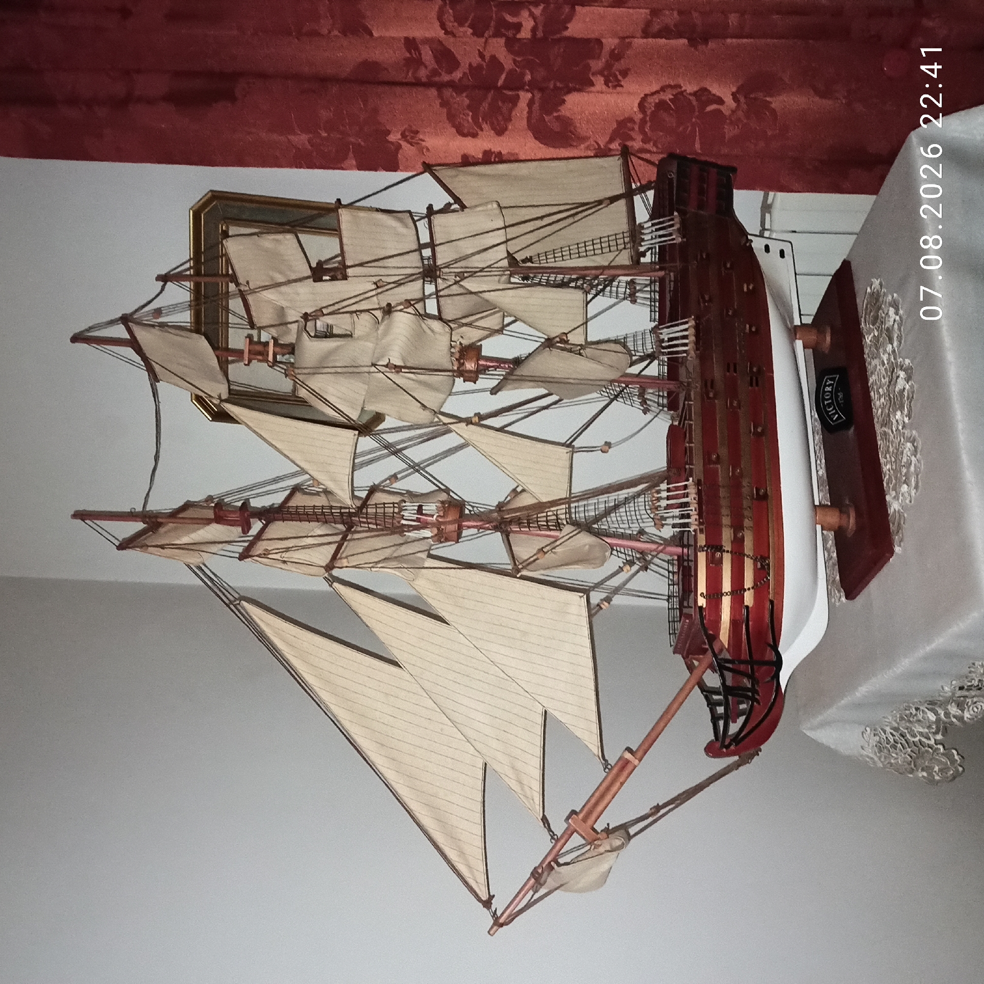 A detailed model of a historic sailing ship, identified by a plaque on its stand as "VICTORY - 1765", is displayed indoors. The ship model is predominantly red and gold with white sails and intricate rigging. It rests on a dark red wooden base supported by two wooden posts, which is situated on a white table covered with a lace tablecloth. The background features a light grey wall and a dark red patterned curtain. The timestamp "07.08.2026 22:41" is overlaid in the lower right corner of the image. There are no people present in the scene. The location in Bitonto, Italy, is not ascertainable from the provided image.
