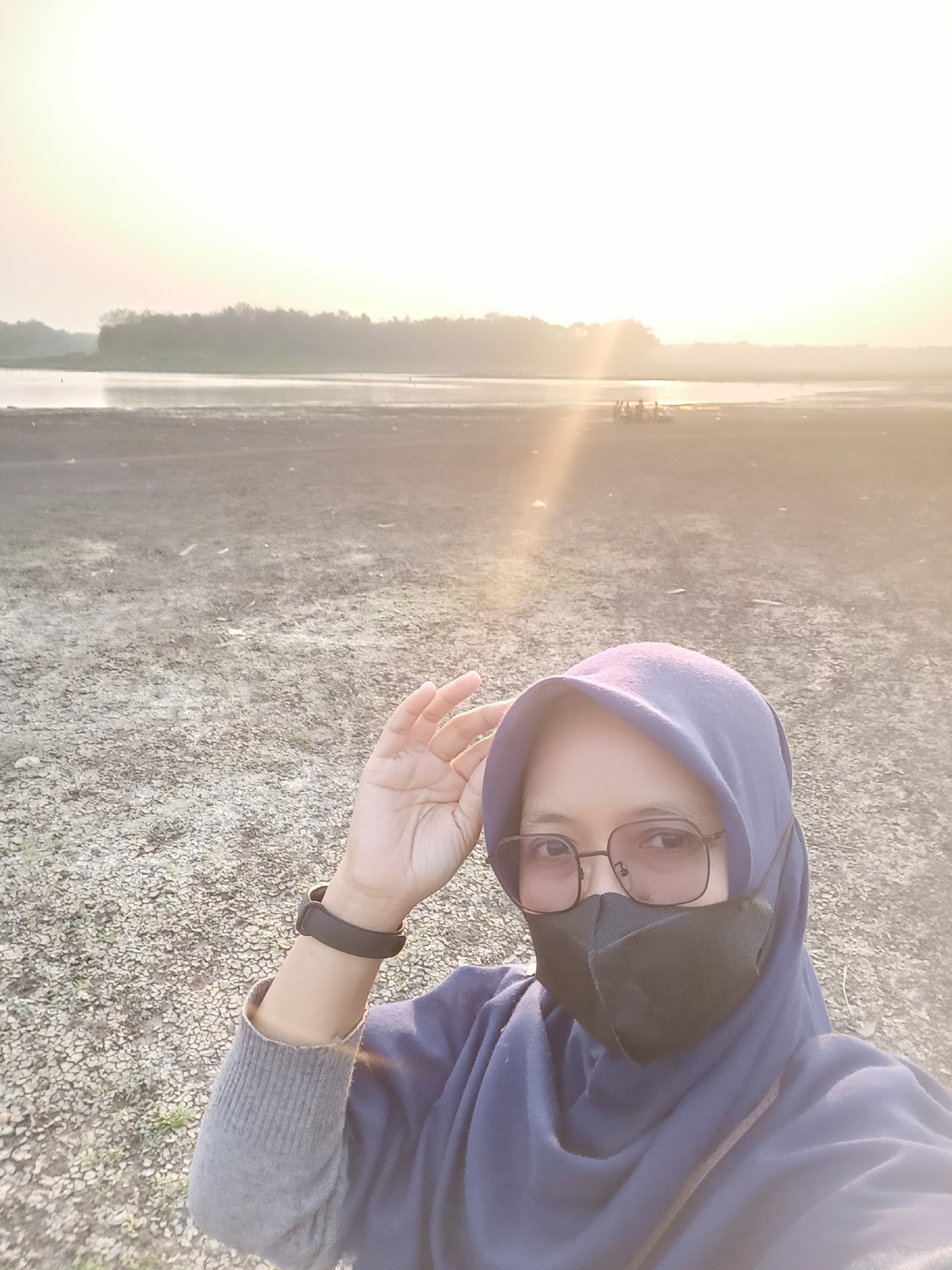 A selfie of a woman wearing a blue hijab, glasses, and a black face mask is taken outdoors. She is standing on a dry, gravelly ground. In the background, there is a body of water with a tree line and a bright, hazy sun setting or rising, casting a warm glow and lens flare over the scene. A few figures appear to be gathered near the water's edge in the distance. The image appears to be taken in the vicinity of the Tanon district in Indonesia.