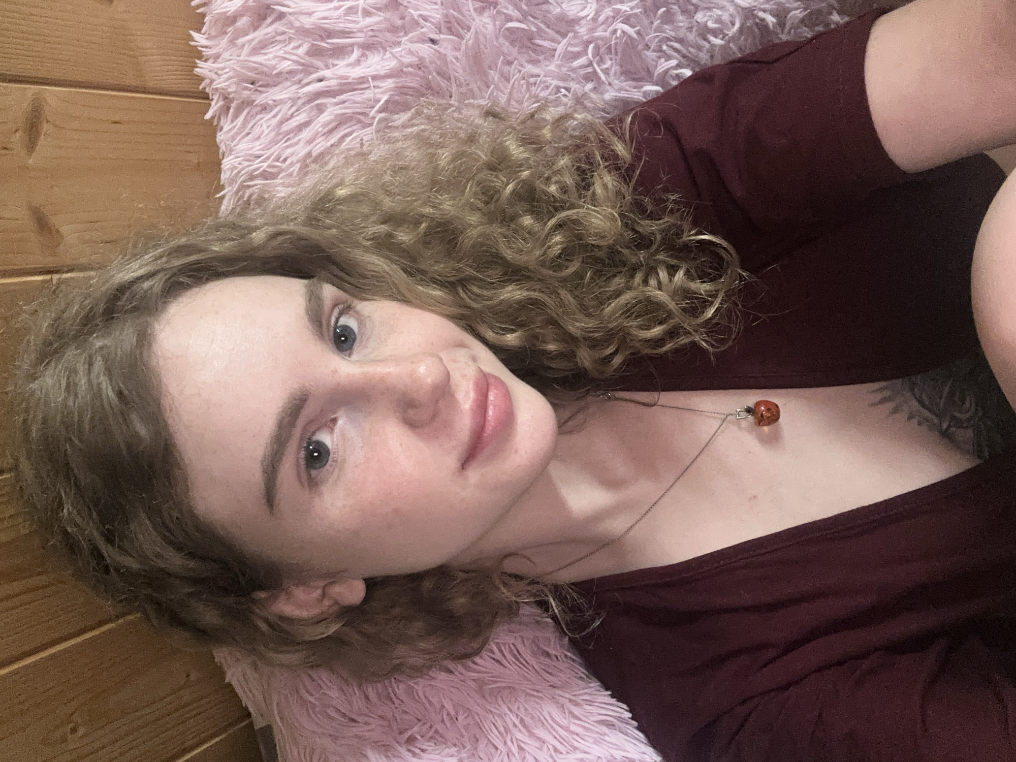 A light-skinned woman with curly blonde hair is lying on a fluffy pink cushion. She is wearing a maroon top and a necklace with a red pendant. There is a wood-paneled wall in the background. The image does not provide sufficient information to determine the woman's location in Vasilyevo, Russia, or to infer any specific actions or interactions.