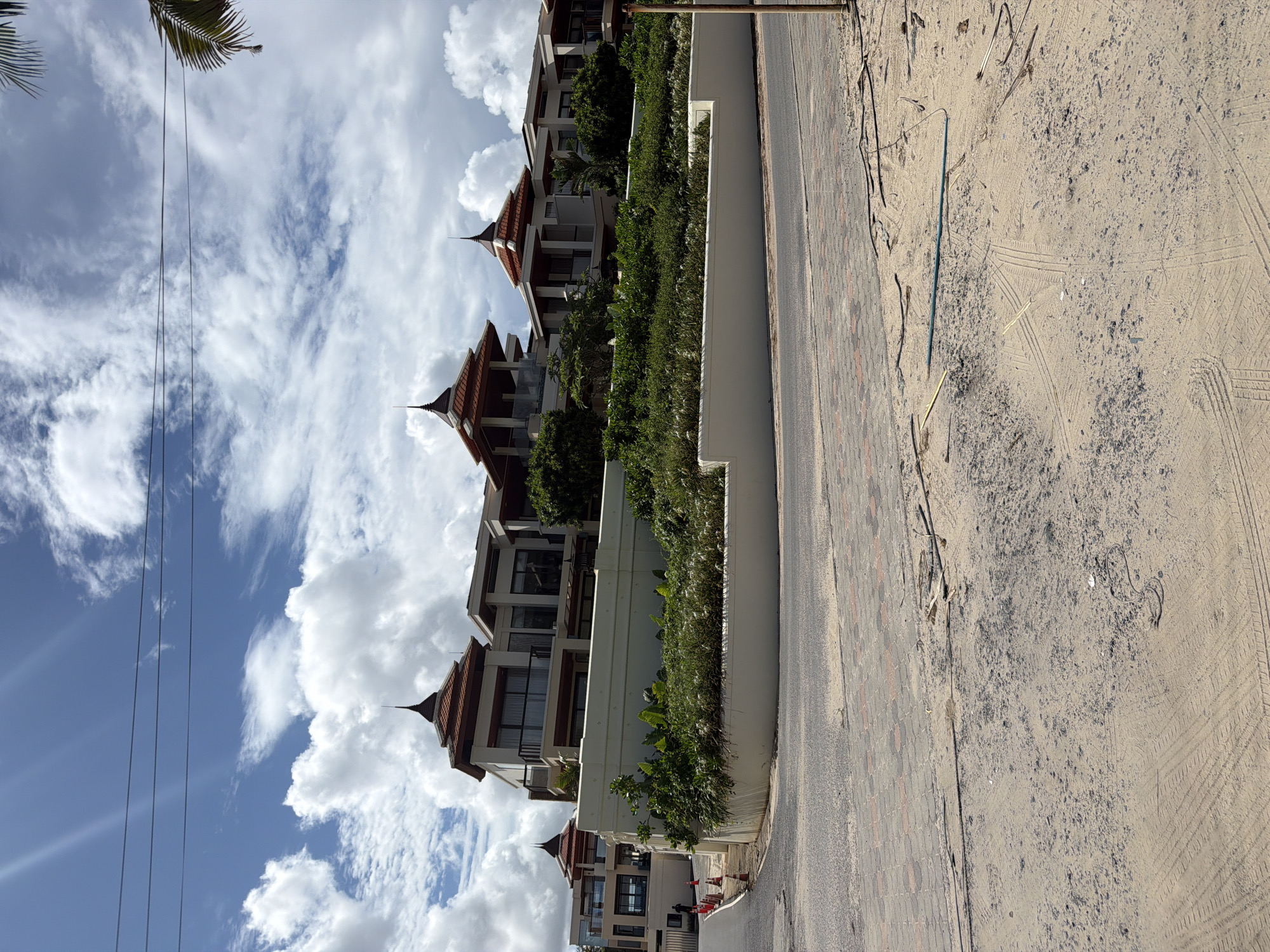 The image shows a row of modern, multi-story buildings with distinctive red, multi-tiered roofs, located in Choeng Thale, Thailand. The architecture features clean lines, large windows, and balconies, suggesting a resort or residential complex. The buildings are situated on a raised embankment covered with lush green vegetation and a white retaining wall. A paved road and a sandy area are visible in the foreground, with some debris and tire tracks on the sand. The sky is partly cloudy with bright sunlight, indicating daytime. The scene captures a well-maintained property in a tropical setting.