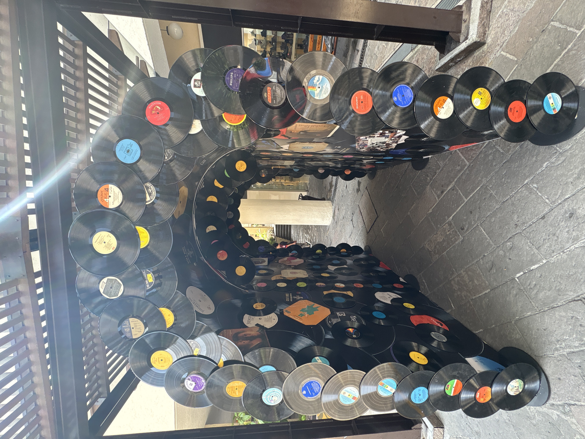 The image depicts an arched entryway constructed from numerous vinyl records, creating a tunnel-like structure. The records are black with various colored labels and are arranged in overlapping circular patterns to form the arch. The ground beneath the arch is paved with stone tiles. Above the arch, a wooden pergola with slatted roofing is visible, and sunlight streams through the gaps, casting diagonal lines of light and shadow. On the right side of the image, a black metal pillar supports the structure. In the background, the tunnel opens into a walkway with buildings and shops on either side, and a person in a red shirt can be seen walking away in the distance. The overall scene suggests an outdoor public space, possibly a plaza or shopping area in the city of Cuernavaca, Mexico.