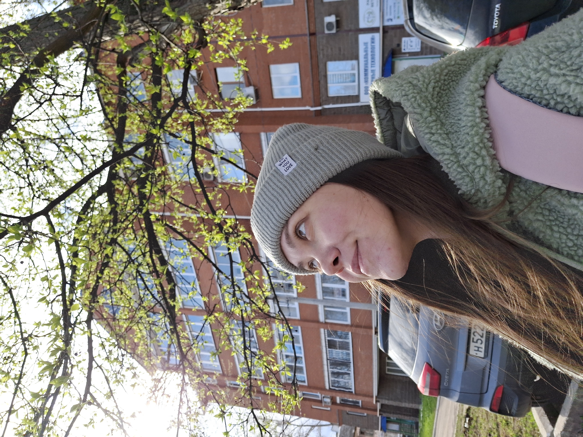 This outdoor image, taken in Chaikovsky, Russia, features a young woman looking upwards and slightly to her left with a subtle smile. She is wearing a grey knitted beanie, which has a white label visible with the text "DO WHAT YOU LOVE." Over a pink top or bag strap, she wears a textured, light green fuzzy jacket. Her long, straight brown hair falls over her shoulders.
The background reveals a multi-story reddish-brown building with numerous windows, some reflecting the bright sky. Several signs are affixed to the building, including one that clearly displays "ООО КОММУНАЛЬНЫЕ ТЕХНОЛОГИИ" (LLC Communal Technologies) in Russian. Parts of parked cars are visible, including a dark grey car identified by "CRETA" on its rear, with a license plate starting "H52", and a black car with "TOYOTA" visible on its back.
Tree branches adorned with fresh, vibrant green leaves frame much of the upper and left side of the scene, indicating spring or early summer. Bright sunlight streams through the leaves, creating some lens flare and suggesting a sunny daytime setting. The overall impression is a casual moment captured in an urban environment.
