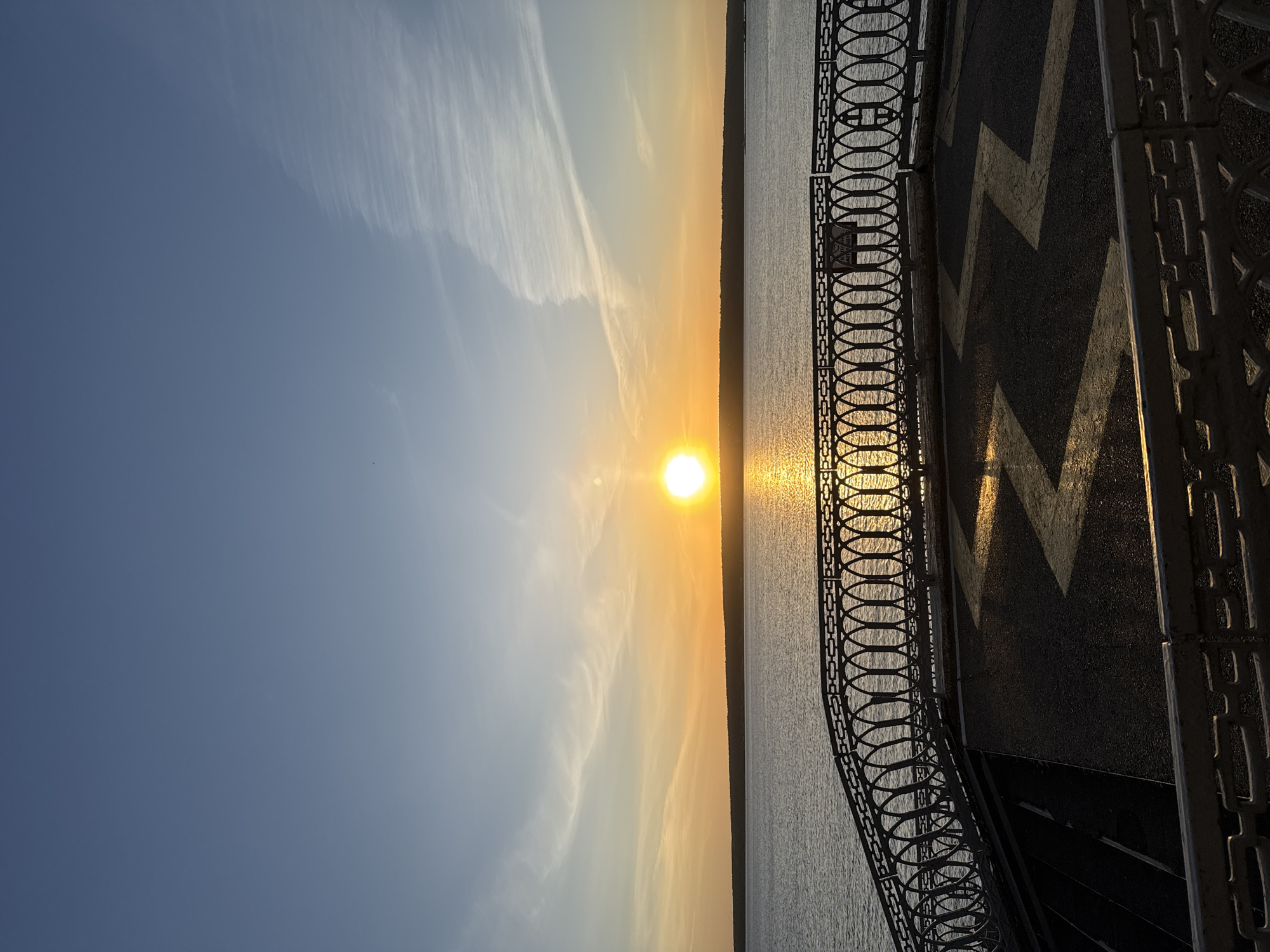 A bright sunset illuminates a body of water with trees visible on the far shore, as viewed from a sidewalk with a decorative metal railing. The sidewalk has a zig-zag pattern. The sky transitions from a clear blue to a golden hue closer to the horizon. The location is Unknown, Unknown.