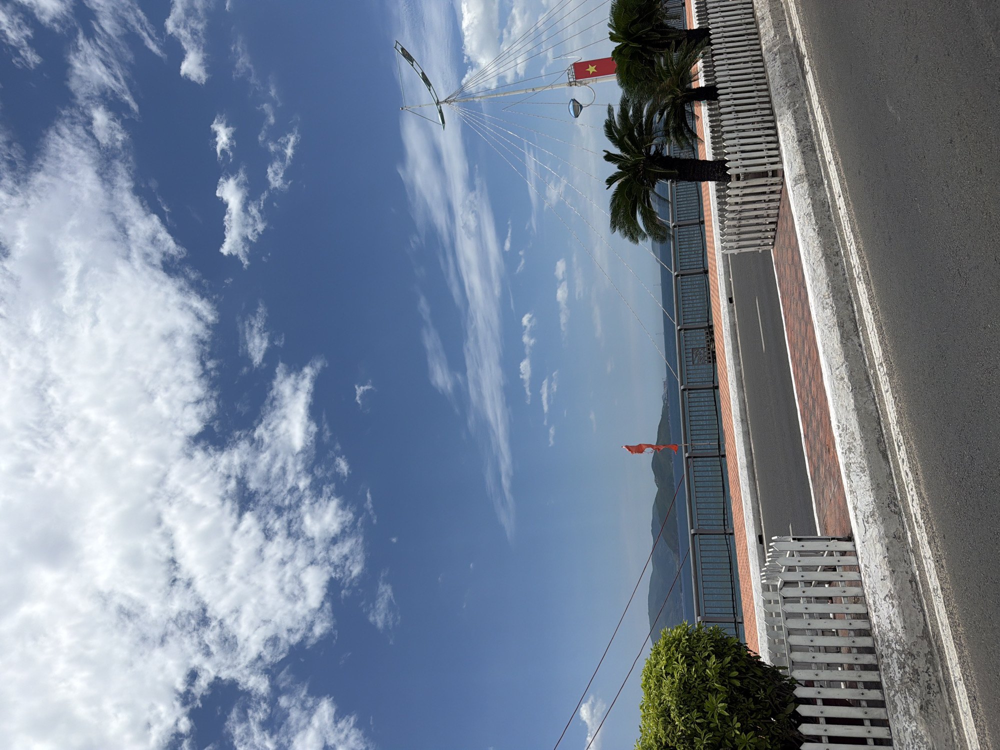The image depicts a street scene in Nha Trang, Vietnam, on a bright, partly cloudy day. The foreground features an asphalt road with white lane markings and a concrete curb. Adjacent to the road, a brick-patterned pedestrian walkway is visible, bordered by a low, white picket fence. Behind the fence, a lush green bush is situated to the left. Further back, a row of palm trees stands against a railing that separates the walkway from a body of water.
In the middle ground, beyond the railing and water, a mountainous coastline stretches across the horizon. A red flag flutters from a pole near the railing. The background is dominated by a vivid blue sky filled with scattered white clouds. To the right, a tall mast with a Vietnamese flag (a red flag with a yellow star) is prominently displayed, suggesting a location near the coast or a waterfront area. The overall impression is one of a scenic, urban coastal environment.
