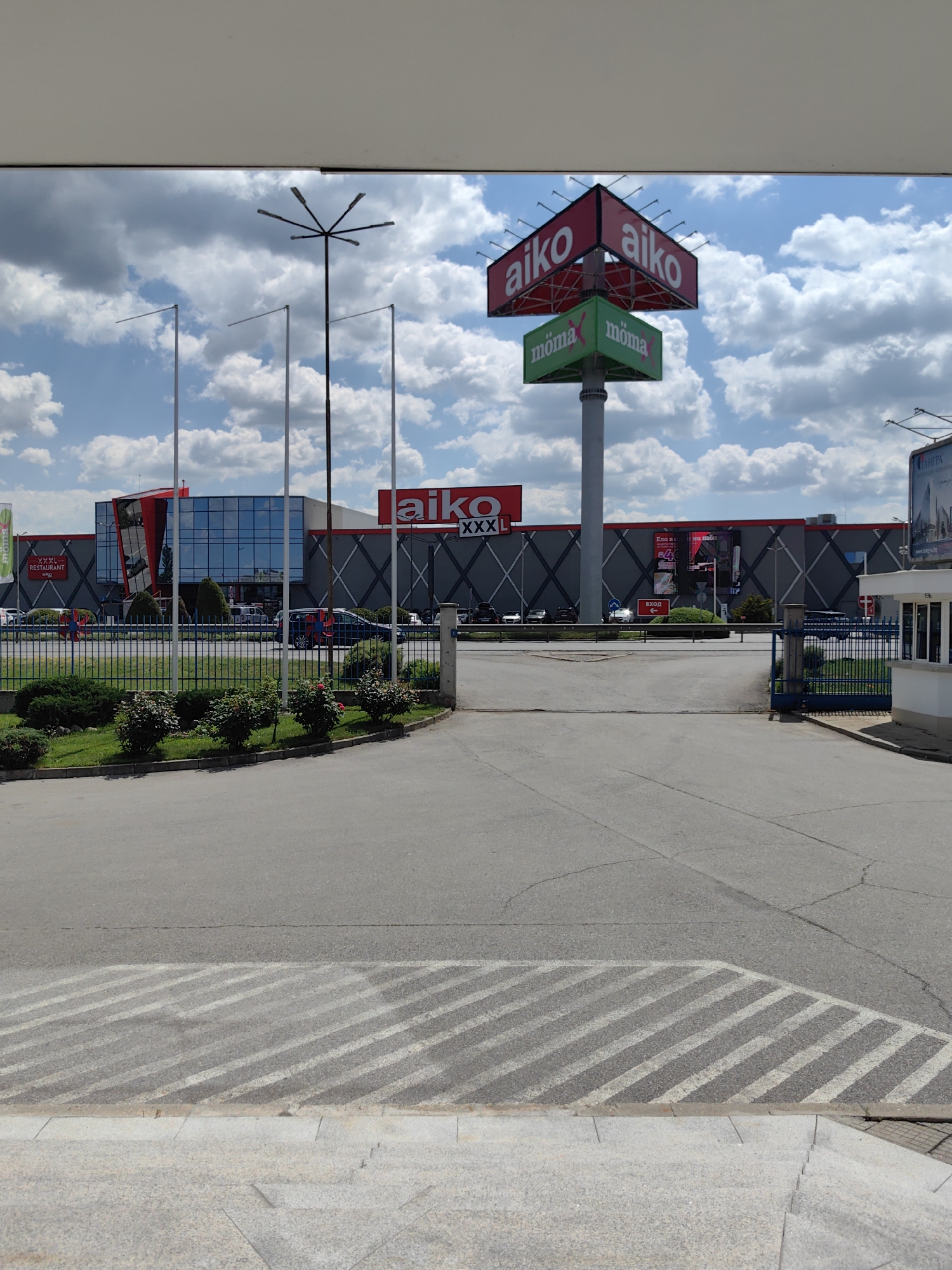 The image captures the entrance of the Aiko XXXL furniture store in Sofia, Bulgaria. The building features a large glass facade and is marked with prominent "Aiko" signs. There is a tall signpost displaying the logos of "Aiko" and "moma". In the foreground, the view is from an elevated position, looking down towards the entrance road, which is marked with striped crossings. There are green hedges and a blue metal fence separating the road from the store's grounds. On the right side, there is a small guardhouse next to the entrance gate. The sky above is partly cloudy. No people are clearly visible in the image. Several cars are parked in the distance.