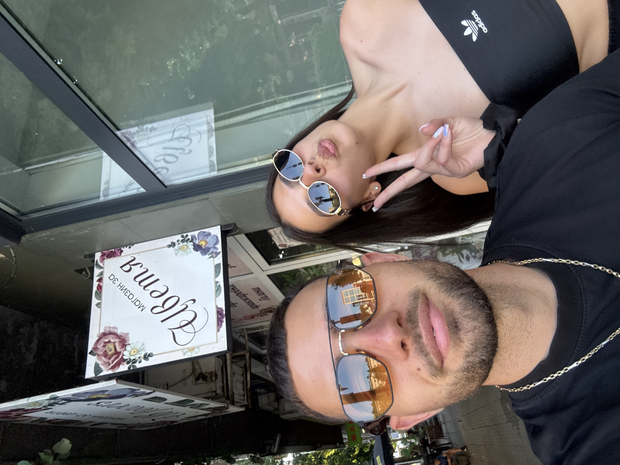 The image captures a selfie of two individuals, a male and a female, outdoors in what appears to be a commercial district. The male, positioned in the foreground on the left, has short dark hair, a light beard, and wears orange-tinted aviator-style sunglasses that reflect a building facade and the sky. He is wearing a black t-shirt and a two-tone silver and gold chain necklace. The female, situated to his right, has long dark hair and wears small, oval, gold-rimmed sunglasses. She is making a peace sign with her left hand, showcasing white-tipped fingernails with a blue accent, and her lips are puckered. She is dressed in a black strapless top featuring a white Adidas logo on the left chest.
Behind the individuals, a prominent square hanging sign for a flower shop is visible. This sign is white with a decorative floral border including a pink peony, white daisies, and purple flowers. The text on the sign is in Cyrillic, reading "МАГАЗИН ЗА ЦВЕТЯ," which translates to "FLOWER SHOP." A second, partially obscured hanging sign, also adorned with floral elements, is visible to the left. Adjacent to the main sign is a large window pane reflecting parts of the shop sign and surrounding trees and greenery. To the far left in the background, an outdoor seating area with a black umbrella and a distant person is discernible. The overall environment suggests a vibrant urban setting under bright daylight. While the Cyrillic text is consistent with the Bulgarian language, specific visual landmarks to identify the precise location as Gabrovo, Bulgaria, are not discernible within the image.