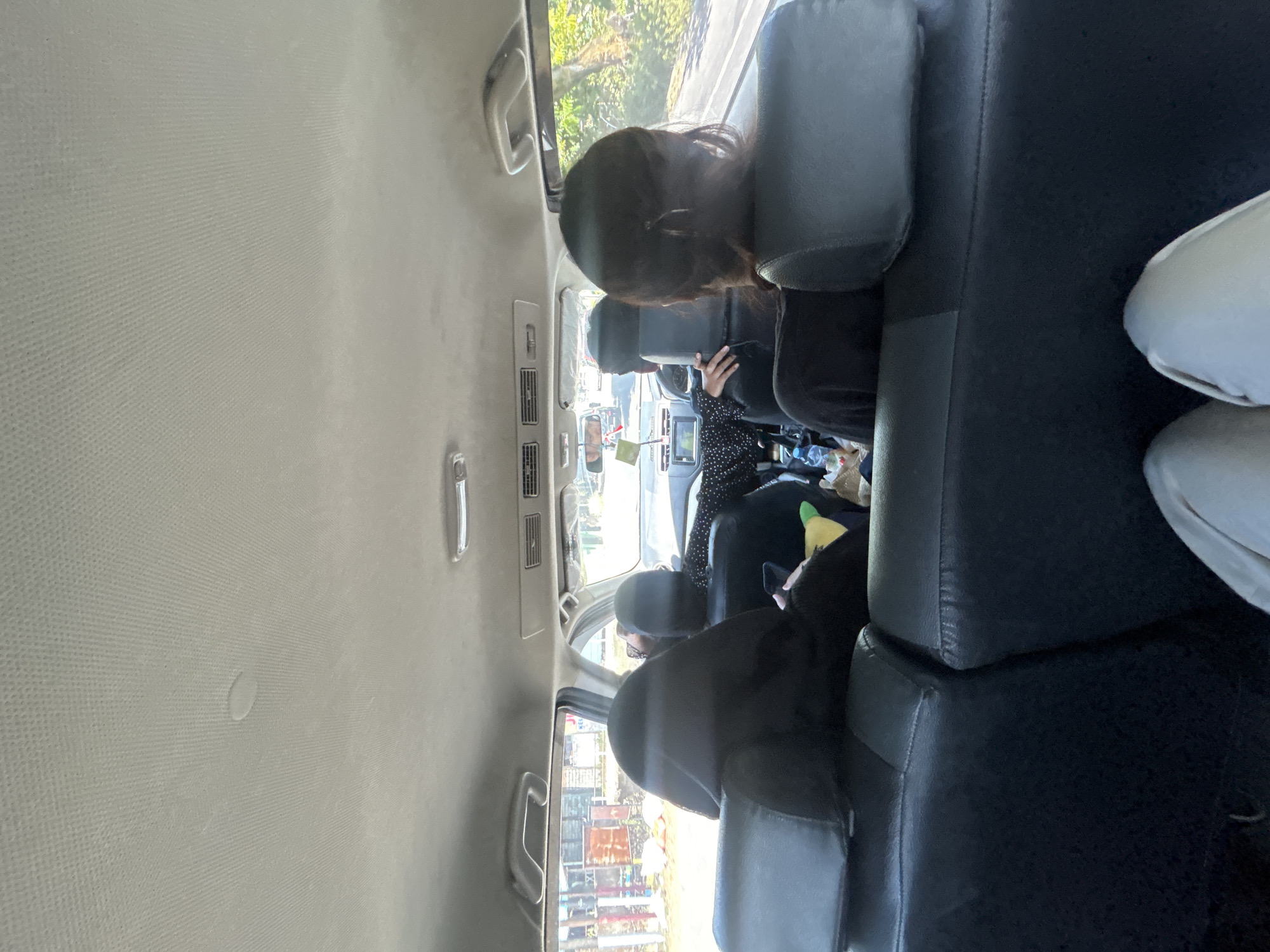 The image shows the interior of a vehicle, likely an SUV, traveling along a road in Kalabbirang, Indonesia. Four individuals are visible. In the foreground, two people are seated in the back row, facing forward. The person on the left wears a dark hooded garment, and the person on the right has dark hair tied back. In the middle row, two individuals are seated. The person on the left, in the driver's seat, appears to be wearing a patterned shirt. The person on the right is looking towards the front and holding a mobile phone. The vehicle's interior includes black leather seats, air conditioning vents on the ceiling, and handles. Outside the vehicle's windows, the scenery suggests an urban or semi-urban environment with buildings and vegetation. The lighting indicates it is daytime.