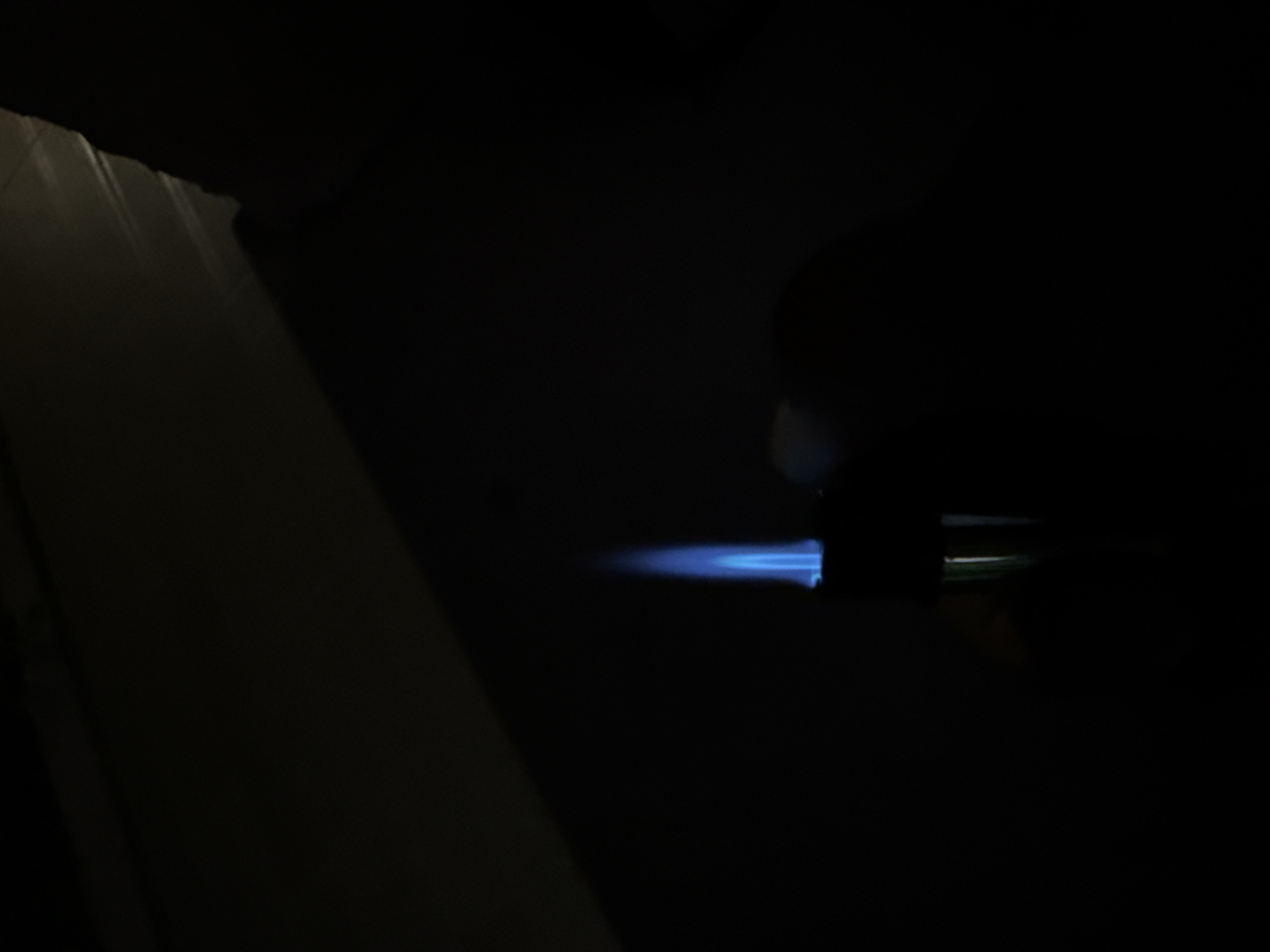 The image depicts a close-up, low-light scene in an undetermined indoor location within the city of Unknown, Unknown. The central subject is a narrow, vertical, bright blue flame approximately 1.5 times taller than its base, emanating upwards from a dark, cylindrical object. This object, likely a torch lighter, has a faint greenish-metallic reflection on its lower right side. To the immediate right of the lighter, an indistinct, dark form consistent with a human hand is present, appearing to hold the device. The background is predominantly dark. In the upper left quadrant, a subtle diagonal edge or surface break is visible against a slightly lighter dark surface, suggesting an architectural feature such as a wall or table edge. The upper right quadrant reveals a textured, dark surface, possibly a wall or draped fabric, with minimal light reflection. The overall environment is characterized by extreme darkness, with the flame serving as the sole significant light source.