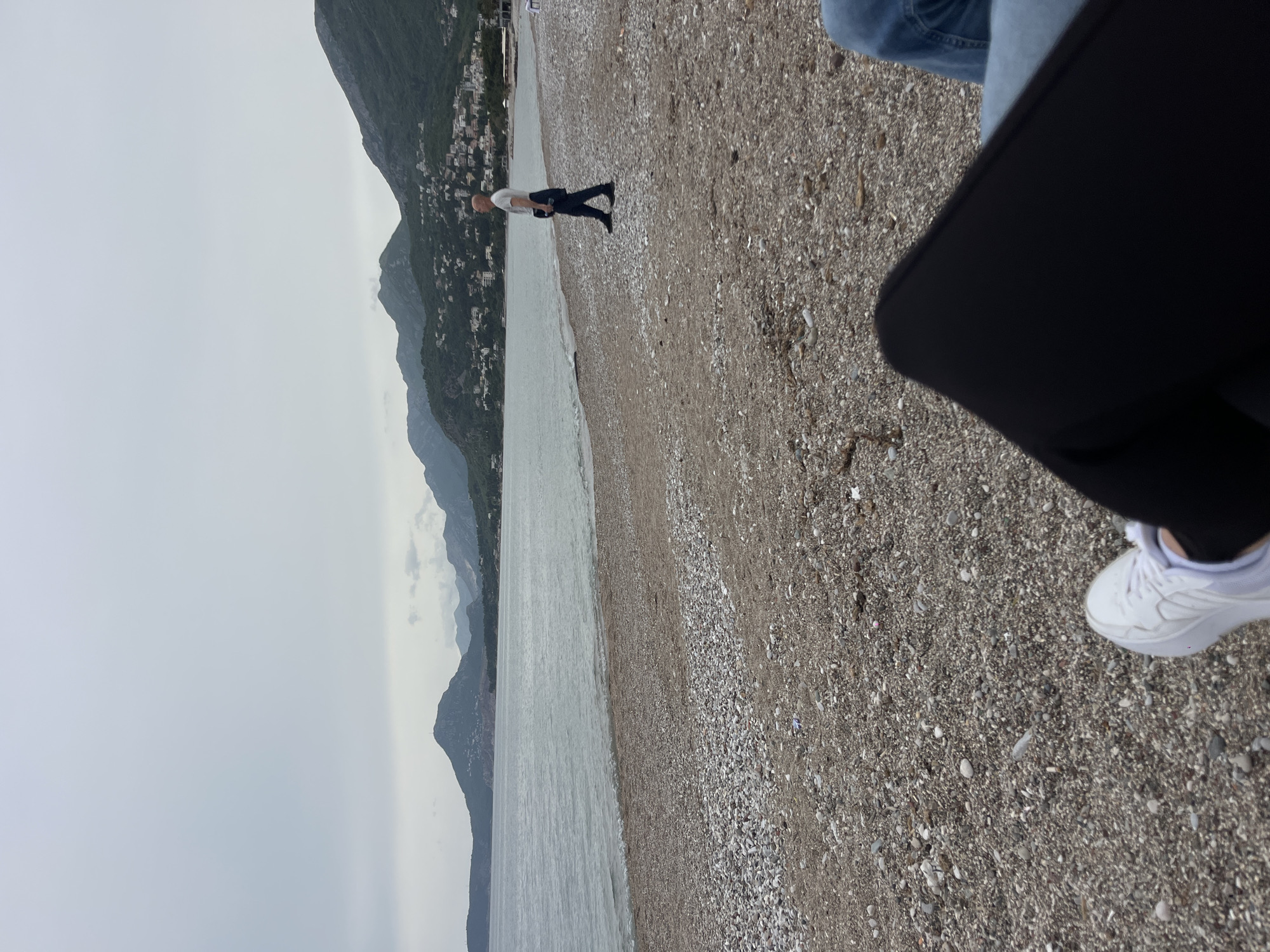 The image depicts a beach landscape with a person walking along the shoreline. In the background, mountains and a town are visible. The foreground shows a pebble beach with a person's legs and white sneaker partially in the frame. The sky is overcast. The town in the background suggests the image was taken near an urban area. Given the location, it is likely the city of Bar, Montenegro.