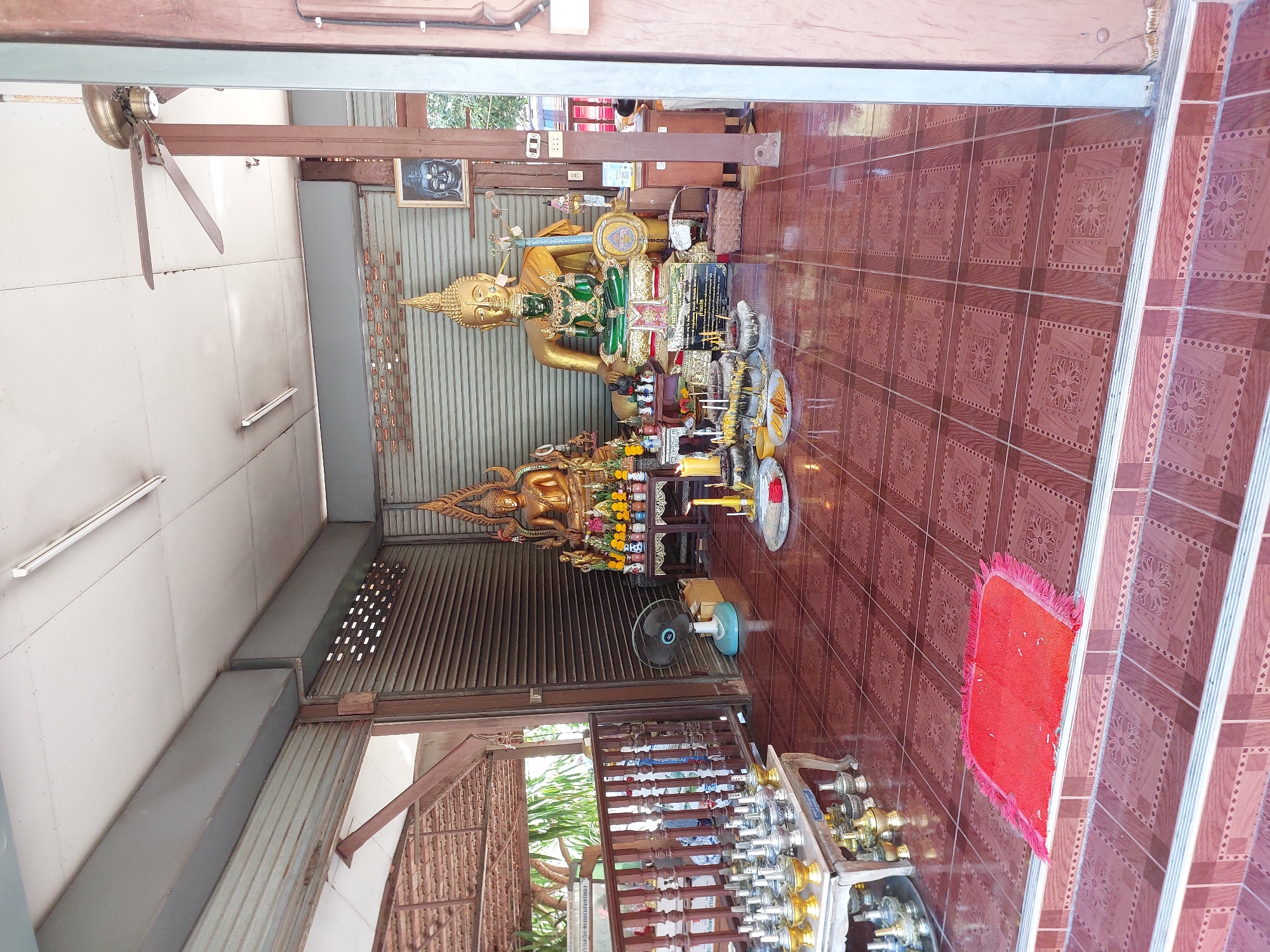 In Baan Plai Laem, Thailand, a shrine area is depicted. Two large, ornate golden Buddha statues are prominently displayed, with a smaller green Buddha statue positioned in front of the larger one. The statues are adorned with offerings, including flowers, incense, and candles. In the foreground, to the left, a table displays numerous silver and gold ceremonial vessels. To the right, a red mat is placed on the floor. The space is open on the left, revealing a wooden railing and foliage beyond. The floor is tiled with a brown, patterned design. A ceiling fan is mounted overhead, and fluorescent lights are visible. The background consists of corrugated metal walls and a roll-up metal door. The scene appears to be an outdoor or semi-enclosed sacred space. There are no people visibly present in the image.