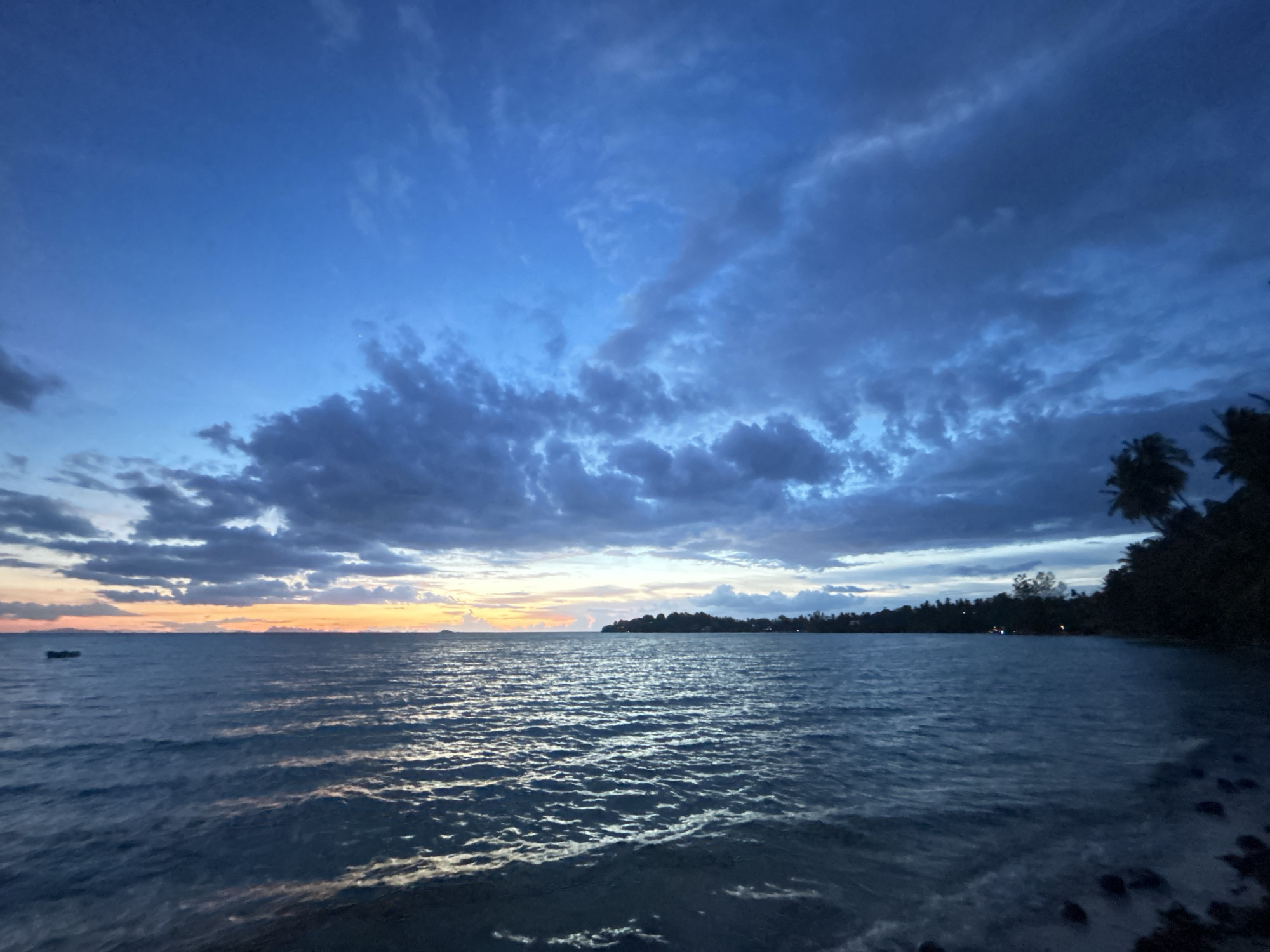 This is a horizontal, eye-level shot of a body of water in the Ko Pha-ngan District, Thailand, at dusk. The sky is a deep blue, with dramatic, dark clouds stretching across it. The sun has just set, and a faint orange and yellow glow is visible along the horizon, peeking through the clouds. The water is calm, with gentle ripples reflecting the fading light. In the distance, a dark silhouette of land is visible, dotted with palm trees and some lights from a settlement. A small, dark boat is also present on the water in the far left of the frame. The foreground shows the edge of the water, with some dark rocks and small waves lapping at the shore. The overall mood is serene and peaceful, characteristic of a tropical sunset.
