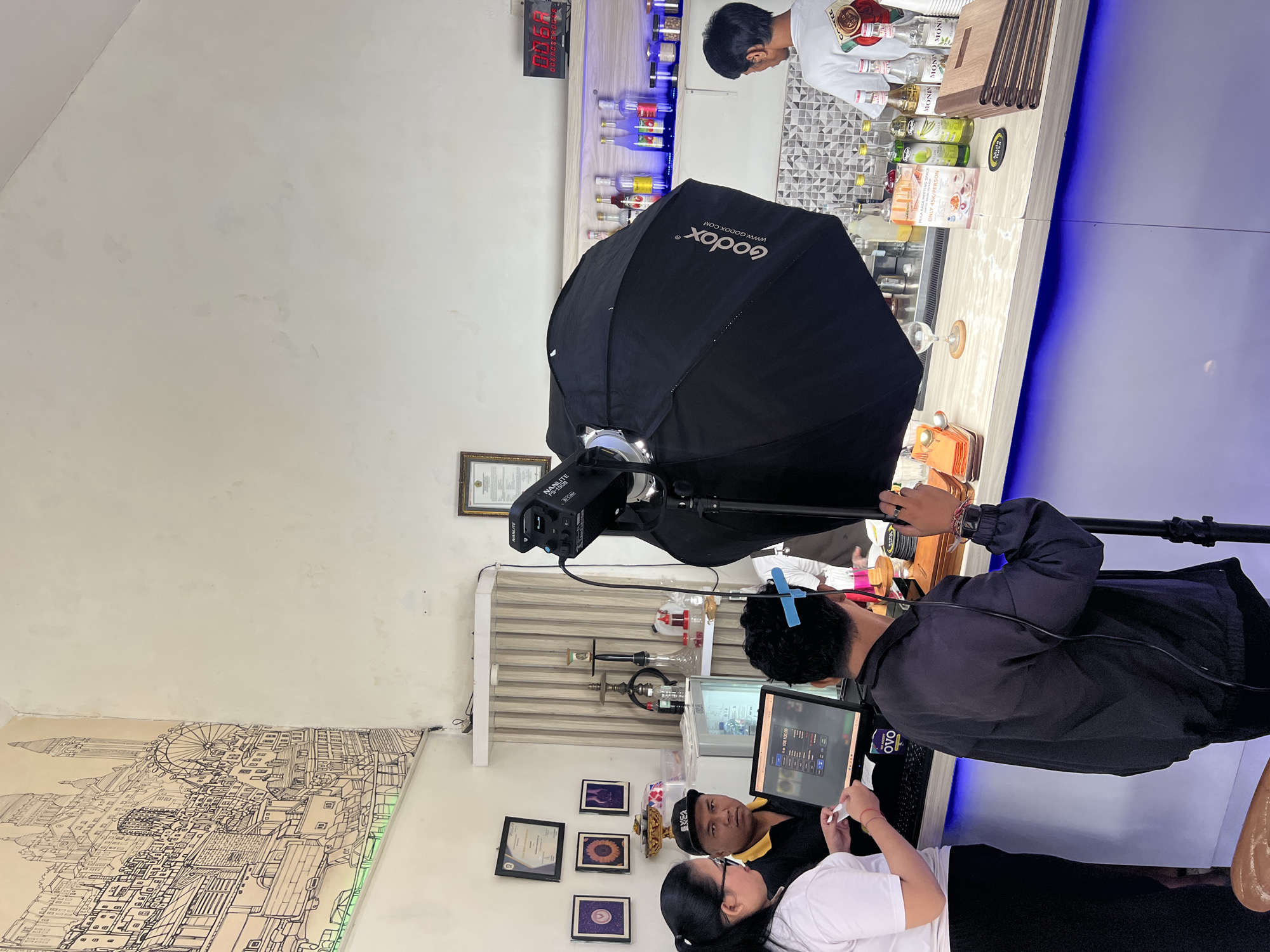 Here is a description of the image:
The image depicts an indoor scene in Sukawati, Indonesia, possibly a business setting. A photography setup dominates the foreground, consisting of a "Nanlite F5-150B" studio light with a "Godox" softbox umbrella attached to a tripod. Three people are visible. A man in a black jacket is adjusting the setup or looking at a laptop. A woman in a white shirt is standing beside him, and another person wearing a black baseball cap and yellow-collared shirt sits behind the laptop.
The setting appears to be a shop or cafe. Behind the photography equipment is a counter with bottles and bar equipment. A person is visible working behind the counter. There's a digital timer on the wall showing "00:68." The wall has decorative elements, including framed certificates/artwork and a sketch of what seems to be a cityscape. There are also signs with the text "MULA RASA HEATING SPACE, ONLY 45K/PERSON" and bottles of "MONIN." The logo "OVO" is visible on the laptop.