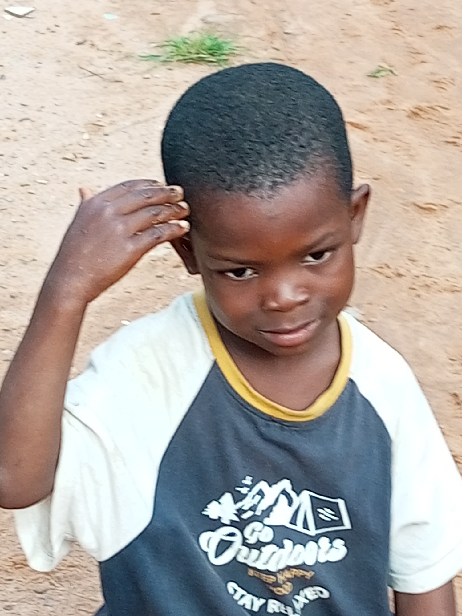 A boy of African descent, appearing to be between 5 and 7 years old, is captured in a medium close-up shot. He has dark, short-cropped hair and is wearing a t-shirt with a graphic design on the front. The t-shirt has a white body with dark gray sleeves and a yellow trim around the neck. The graphic on the t-shirt appears to depict mountains and tents, with text that reads "Go Outdoors" and "Stay Relaxed". The boy is looking towards the viewer's right with a somewhat serious expression. His right hand is raised and touching his head, near his ear. The background is out of focus and appears to be a sandy or earthy surface with some sparse vegetation. The lighting suggests an outdoor environment, possibly during the day. This image is likely from Tempure, Nigeria, given the context.