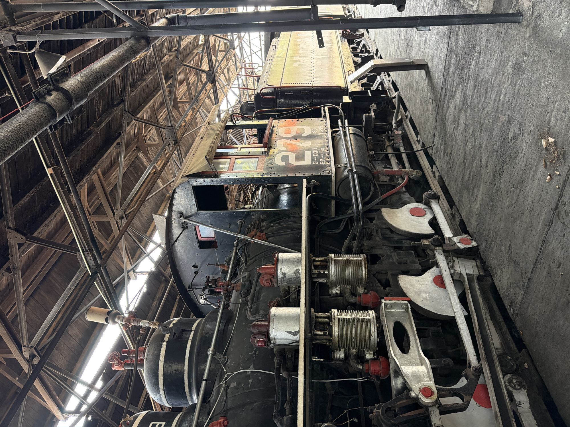 A steam locomotive numbered "293" dominates the frame. It is parked indoors in a building with a visible wooden framework. The walls of the building are concrete, marked with dirt. The location is likely a museum or historical site in Cuautla, Mexico, given that locomotive 293 is located in that city. The locomotive's machinery, including cylinders, valves, and pipes, are prominent. No people are visible.