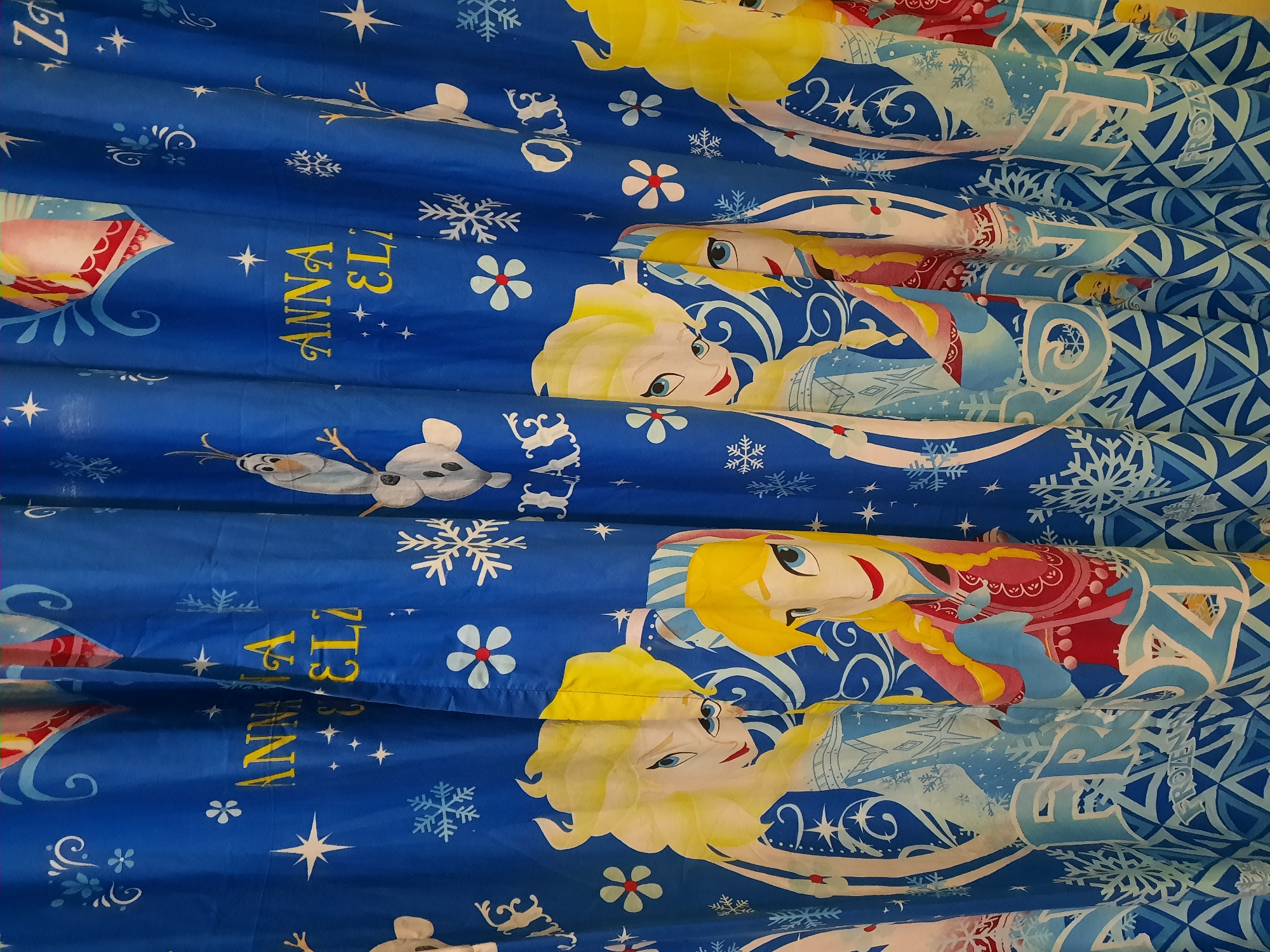 The image shows a close-up, head-on view of a blue shower curtain featuring characters and designs from the Disney movie "Frozen." The curtain is adorned with images of Elsa and Anna, as well as Olaf the snowman. Snowflakes and stars are scattered across the blue fabric, adding to the winter theme. The text "ANNA" and "ELSA" are prominently displayed in yellow letters, partially obscured by the folds of the curtain. The bottom of the curtain reveals a pattern of blue geometric shapes and the word "FROZEN" in large, stylized white letters. The lighting suggests an indoor setting, likely a bathroom. The curtain is hanging, indicating it is in use. There are no people visible in the frame. The image does not contain any information about its specific location in the city of Lhokseumawe, Indonesia.