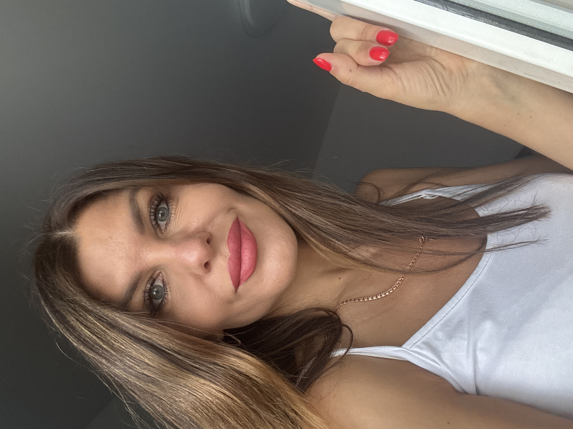 A woman with brown hair, fair skin, and wearing a white tank top is posing for a close-up selfie indoors. She has applied red lipstick and nail polish. Her right hand is visible, holding onto a white window frame. The background consists of a dark gray wall. The location is presumed to be in Chaikovsky, Russia, based on the prompt.