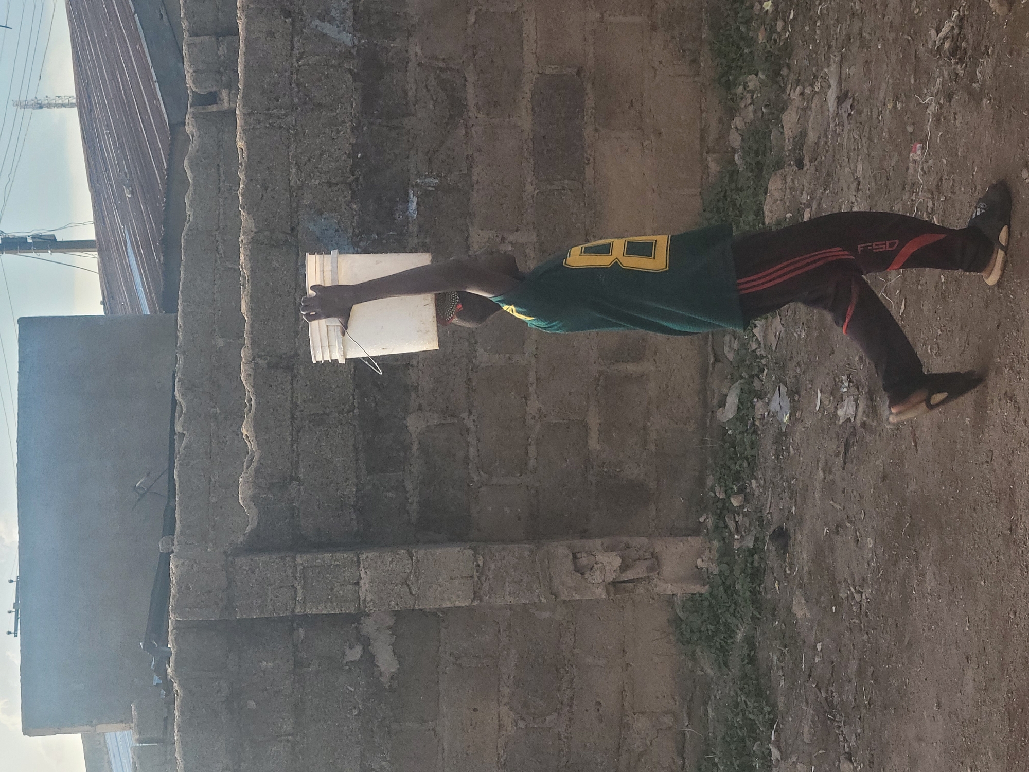 A vertical shot, rotated 90 degrees clockwise, captures a person holding a white bucket outdoors. The individual, appearing male, stands with arms extended, holding the white plastic bucket directly in front of them with both hands. They are wearing a dark green short-sleeved t-shirt with a prominent yellow letter 'B' (possibly part of 'B8' or 'BD') on the back. Their lower body is clad in dark-colored trousers featuring three red stripes down the side and the text "FSD" visible on one leg. Light-colored sandals are on their feet.
The background consists of a textured wall made of grey concrete blocks, extending across the mid-ground. Beyond the wall, a corrugated metal roof is visible, along with utility poles and power lines against a bright, clear sky. The ground in the foreground and to the person's right is uneven dirt, scattered with small bits of debris and patches of sparse green vegetation. The lighting suggests it is daytime, under clear weather conditions. The overall scene, including the building materials and ground cover, aligns with the provided context of Jos, Nigeria.