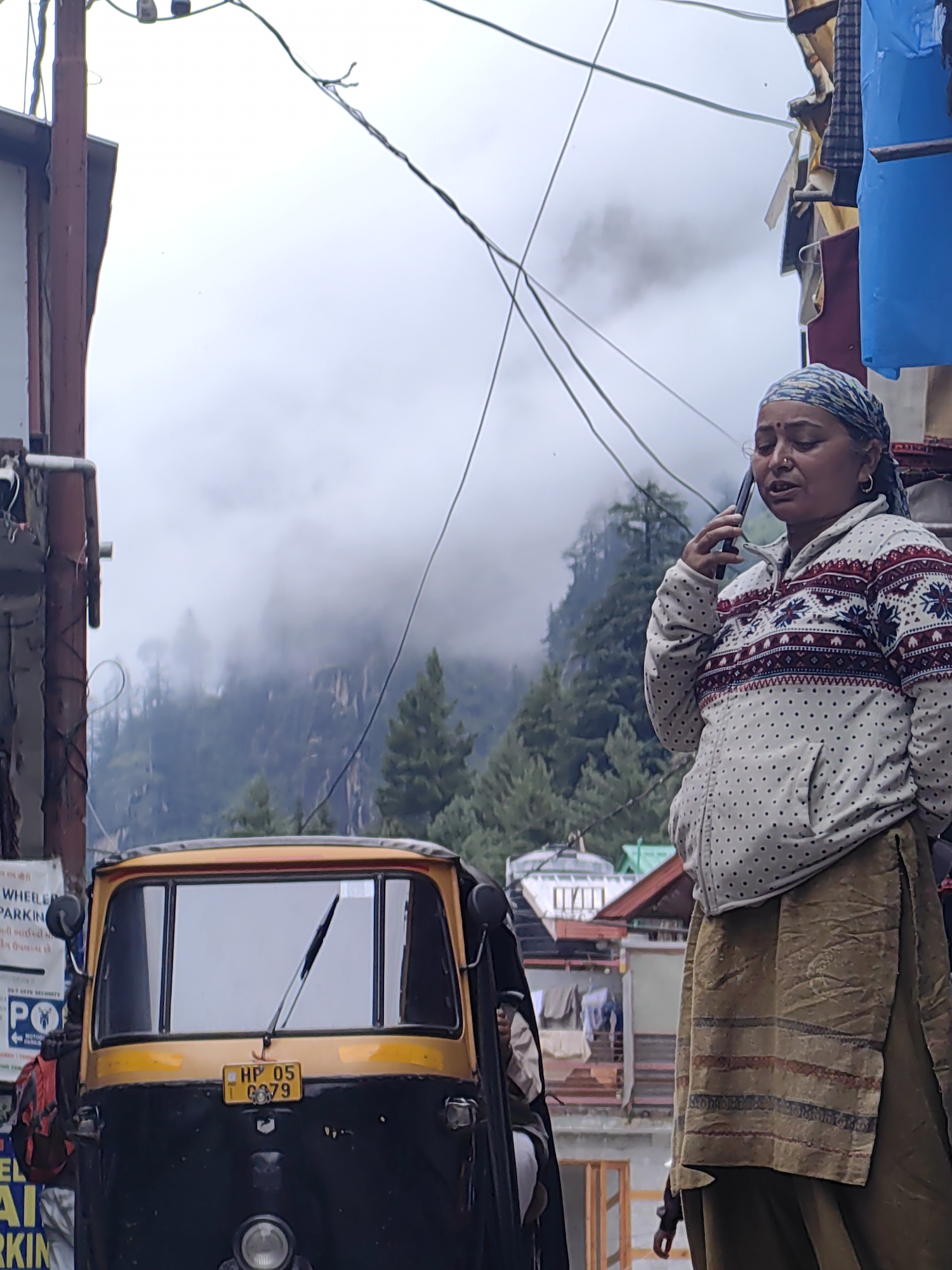 In Vashisht, India, a woman with a blue and white headscarf and a patterned sweater is speaking on a mobile phone. She has a bindi on her forehead and is wearing a green skirt. In the foreground, the front of a yellow and black auto-rickshaw is visible, with a license plate reading "HP 05 G379". Behind the woman, a hazy view of forested mountains and buildings is present, with power lines crisscrossing the sky.