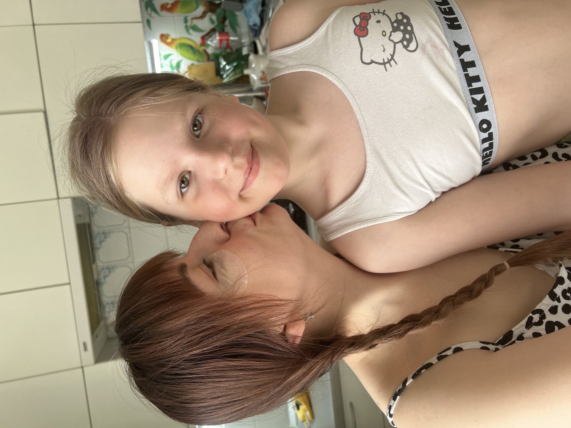 This image captures a tender moment between an adult and a child, seen from a high angle looking down. The image is rotated 90 degrees to the left.
In the foreground, an adult with reddish-brown hair is seen from the side, positioned below the child. Her hair is styled in a long, single braid that extends down her back. She is wearing a black and white spotted garment, which appears to be a bra or bikini top. A transparent, possibly gel-like, under-eye patch is visible beneath her left eye. Her face is turned towards the child, and her lips are pressed gently against the child's forehead, suggesting a kiss. An earring, possibly a small stud or simple drop, is visible in her left ear.
Above and slightly to the right of the adult is a young child, who is looking directly at the camera with a gentle smile. The child has light brown hair, parted slightly to the left, and bright, warm brown eyes. She is wearing a white tank top or sports bra. On the front of the top, the iconic cartoon character "Hello Kitty" with a red bow is clearly visible. The elastic band at the bottom of her top also features the text "HELLO KITTY" in black letters on a grey background.
The background primarily consists of tiled walls. To the left and behind the adult, there are white square tiles. To the right and behind the child, the tiles are more decorative, featuring a pattern with green and yellow parrots on branches against a light background, possibly indicating a kitchen or dining area. Various items like bottles and containers can be partially seen in the blurred background. The overall impression is one of affection and closeness between the two individuals.