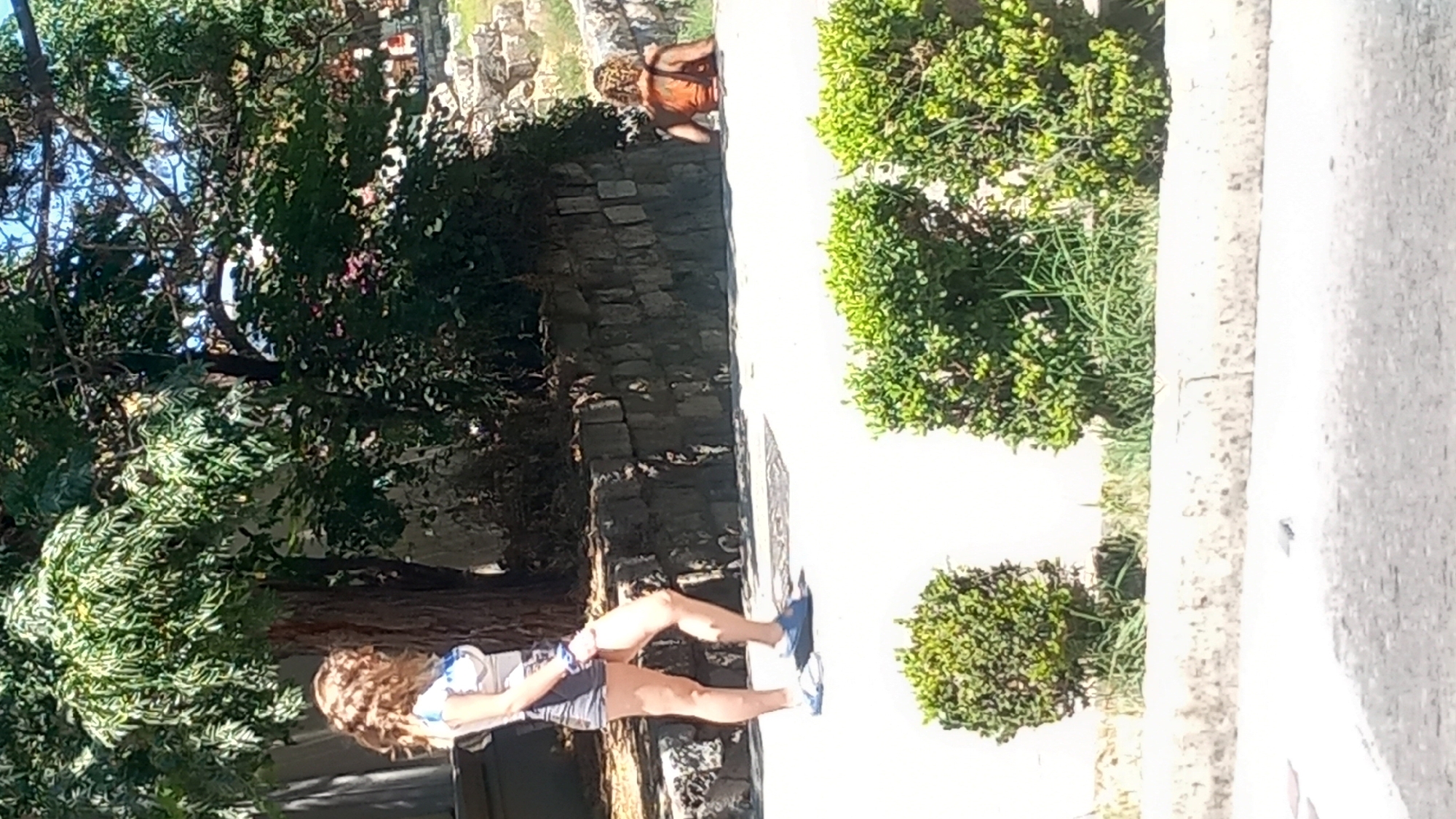 In the city of Kos, Greece, a woman with wavy blonde hair is walking on a paved path. She is wearing a white and blue top and grey shorts. To her right, a person with blonde hair is sitting in a body of water, facing away from the camera. The background features lush green trees, a stone wall, and a rocky embankment. The scene is illuminated by bright sunlight, casting shadows on the ground.