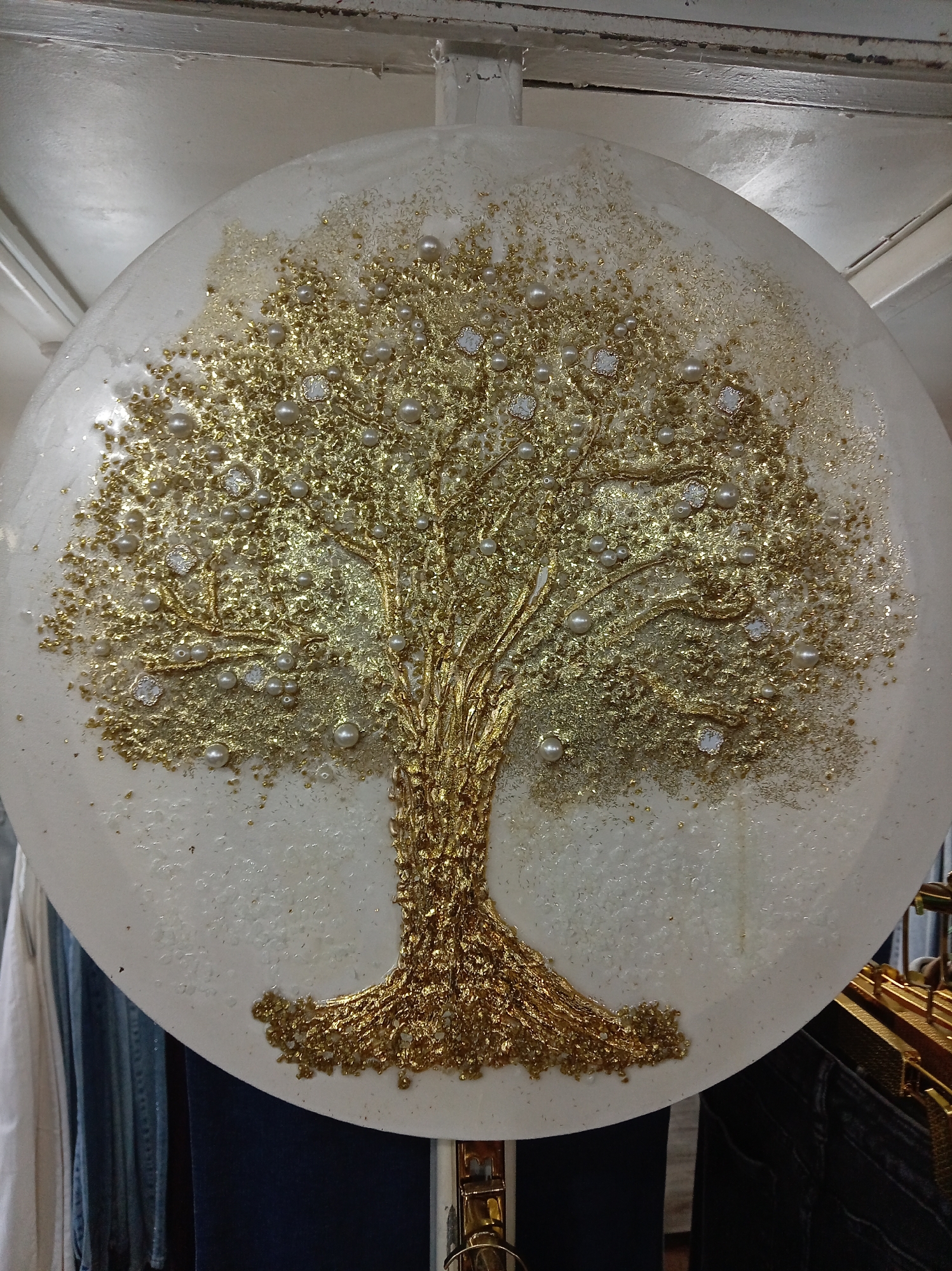The image shows a close-up of a decorative, circular artwork featuring a stylized golden tree. The tree is rendered with a textured, raised surface, appearing to be made of a golden material that mimics bark and branches. Embedded within and around the tree's foliage are numerous small, pearl-like beads and clear, faceted stones that sparkle, suggesting they might be crystals or rhinestones. The background of the artwork is a textured white or off-white surface, with some areas appearing to have a granular or glittery finish. The artwork is circular and appears to be displayed against a backdrop of clothing and hangers, suggesting it is in a retail environment or a personal display. There is no discernible location in the city of Donetsk, Ukraine, indicated within the image itself.