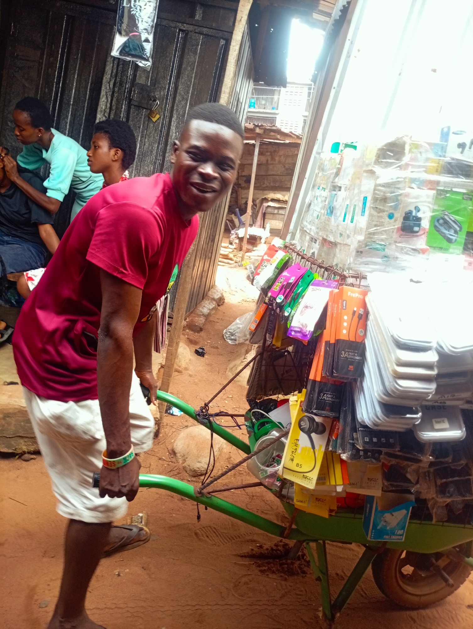 A man, wearing a red t-shirt and white shorts, smiles as he pushes a green wheelbarrow laden with cell phone accessories. The wheelbarrow is filled with various items such as phone cases, chargers, and headphones. In the background, three other people are visible, and the scene appears to be an outdoor market stall. The ground is sandy, and the structures in the background suggest a developing urban environment.