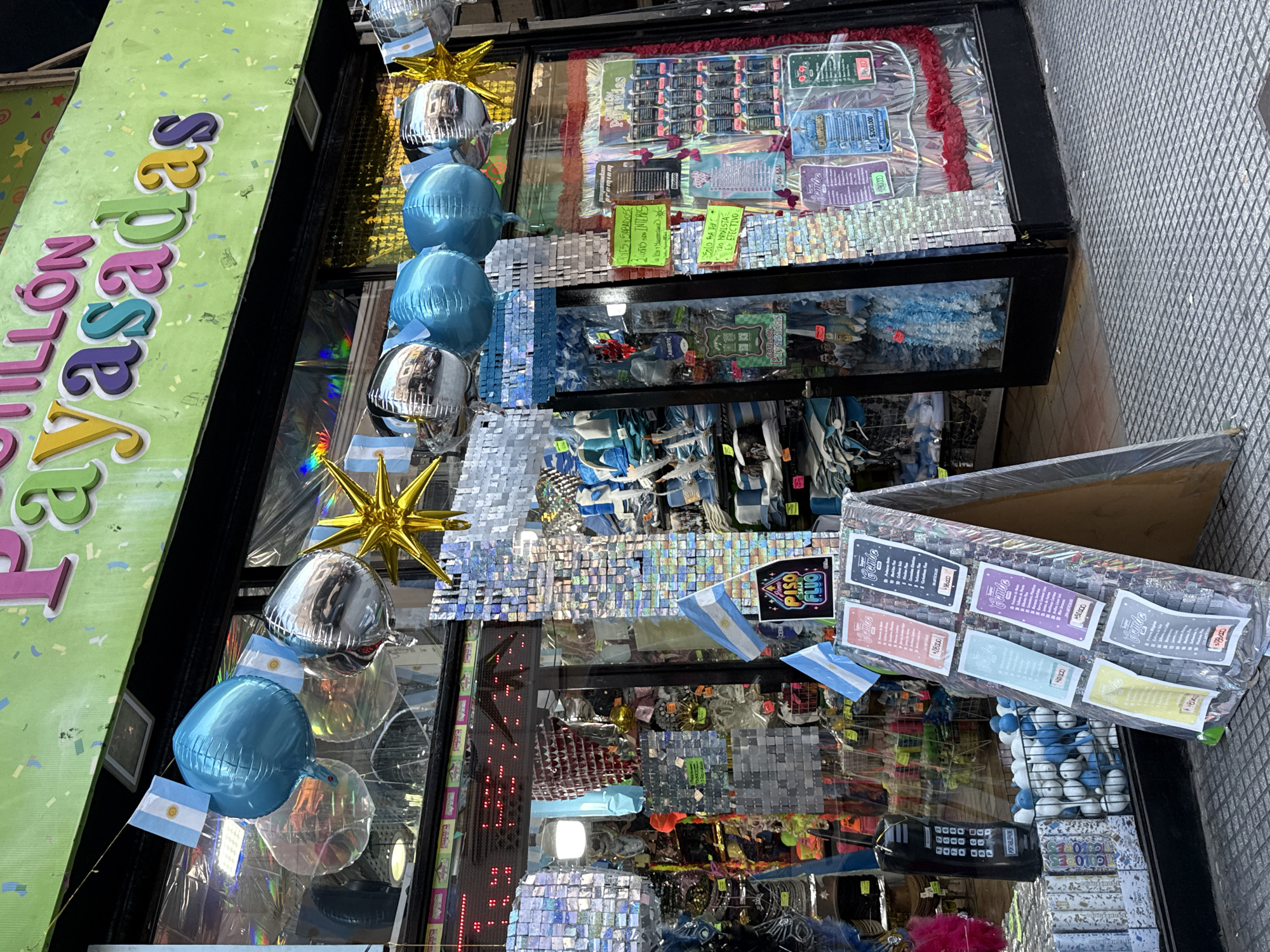 The image captures a vibrant storefront, likely a party supply or novelty shop named "COTILLÓN Payasadas," located in Buenos Aires, Argentina. The prominent green sign on the left features the store's name in colorful, raised letters, indicating a shop specializing in party articles and joke items.
The large display windows are filled with a vibrant assortment of merchandise and decorations, indicative of a celebratory or festive theme. Numerous blue and silver foil balloons are interspersed with small Argentinian flags, a strong visual cue for the country. A gold star-shaped balloon adds to the festive decor.
The displays showcase a variety of items. One section features scratch-off cards or lottery tickets, some labeled "Campeones" (Champions) and showing monetary values, possibly referencing a recent victory (like the World Cup). Below these, reflective, square-shaped ornaments create a sparkling backdrop for other products. Further items include multiple packets labeled "Crembo" with prices in Argentine Pesos (e.g., "$2000," "$18000"), a black toy "PORTABLEPHONE," and a box containing small blue and white spherical items.
Visible text on various signs includes "ABIERTO" (Open), indications for "Cuotas sin interés" (installments without interest), and "PISO FLUO" (Fluorescent Floor). A sign also mentions a discount for cash ("EN EFECTIVO"), and prices up to "$200.000" are displayed. The abundant use of Argentinian flags throughout the decorations firmly places the scene in Argentina.
The bright natural light suggests it is daytime. No people are visible, and the scene is a colorful, densely packed retail display.