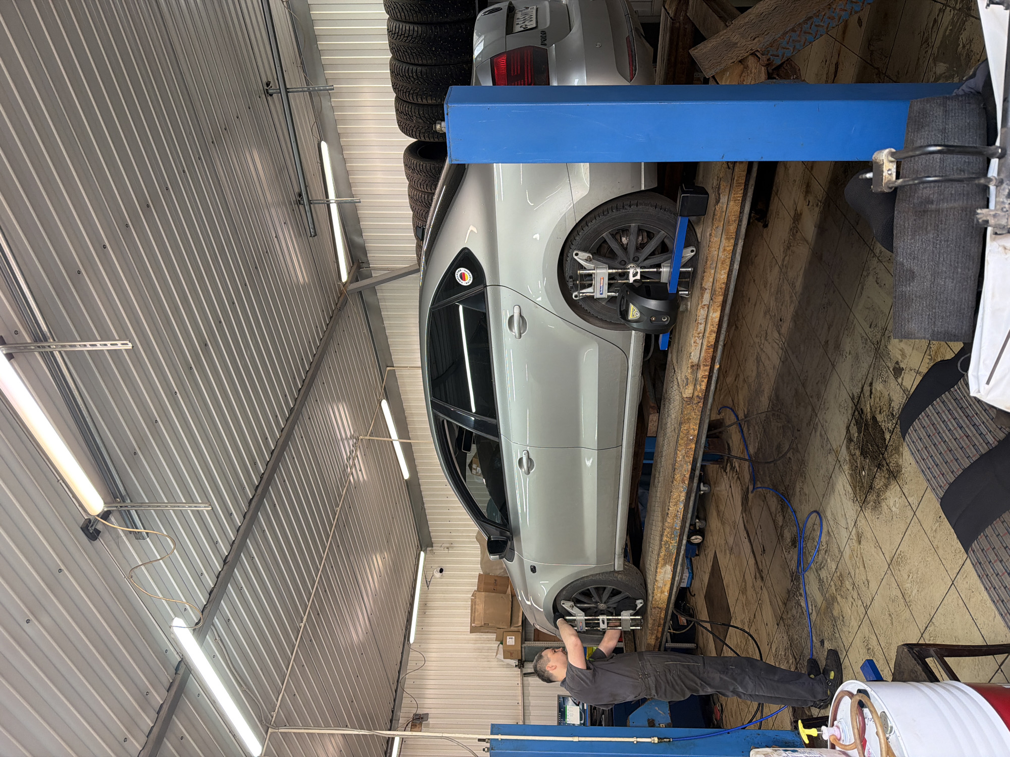 A man in a dark grey mechanic's jumpsuit is standing next to a silver sedan that is raised on a hydraulic lift in what appears to be a garage or workshop. The man is holding a device and adjusting it near the front wheel of the car. Two other wheels of the car have similar devices attached to them, which are connected to a computer console to the left of the man. The garage has a corrugated metal ceiling with fluorescent lights and white paneled walls. Stacked tires are visible in the background. The floor is tiled and appears wet in places, with oil stains. A large white barrel with red and blue accents is to the left of the man, and a portion of a car seat is visible in the foreground. The license plate on the car is partially visible and appears to be Russian. The setting is a car repair shop in Izhevsk, Russia.