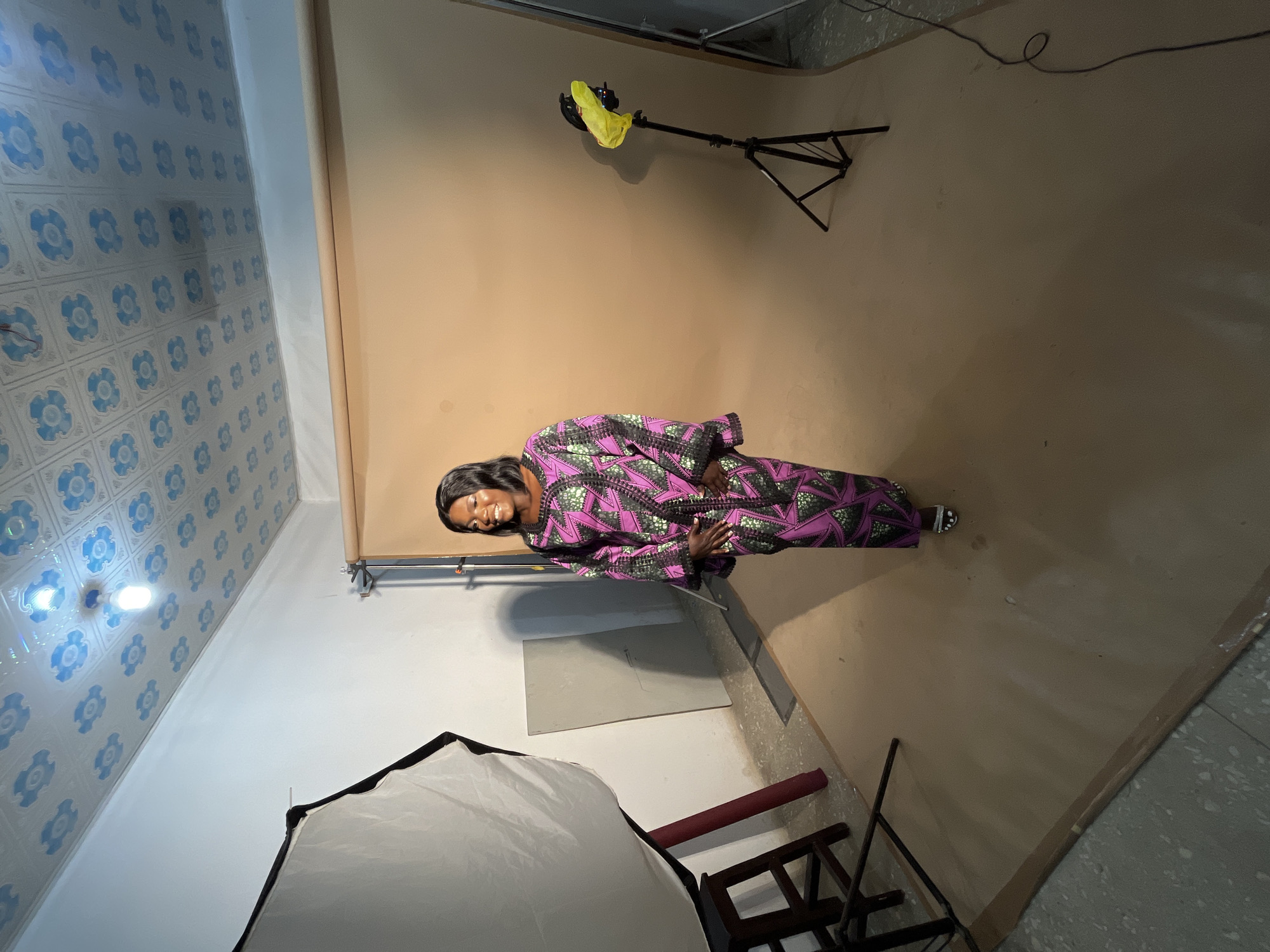 A full-body medium shot captures a Black woman standing in what appears to be a photography studio in Gura Top, Nigeria. She is positioned in the center of the frame, facing slightly to her left, with a warm smile. The woman is wearing a vibrantly patterned long dress in shades of pink, green, and black, with a geometric design. Her hair is dark and styled in a bob, framing her smiling face.
The background consists of a large, seamless tan backdrop that covers the wall and floor. To the left of the woman, a large white softbox light modifier is visible, supported by a dark tripod and red pole. A dark wooden stool is positioned in front of the softbox. To the right, another tripod stands, holding a small light with a yellow plastic bag over it. The floor in the foreground is a mixture of grey concrete and the tan backdrop material, with some taped seams. The ceiling is covered with white tiles featuring a repeating blue floral pattern, and a single light bulb hangs down. The overall impression is of a simple, functional studio setup.